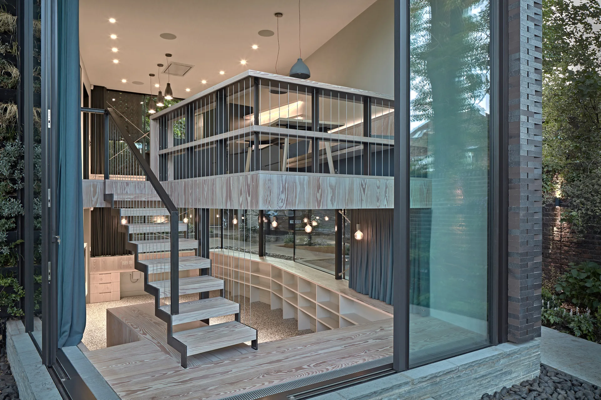 A modern multi-level interior featuring an open-tread wooden staircase, a mezzanine with cable railings, and a large glass facade overlooking a garden.