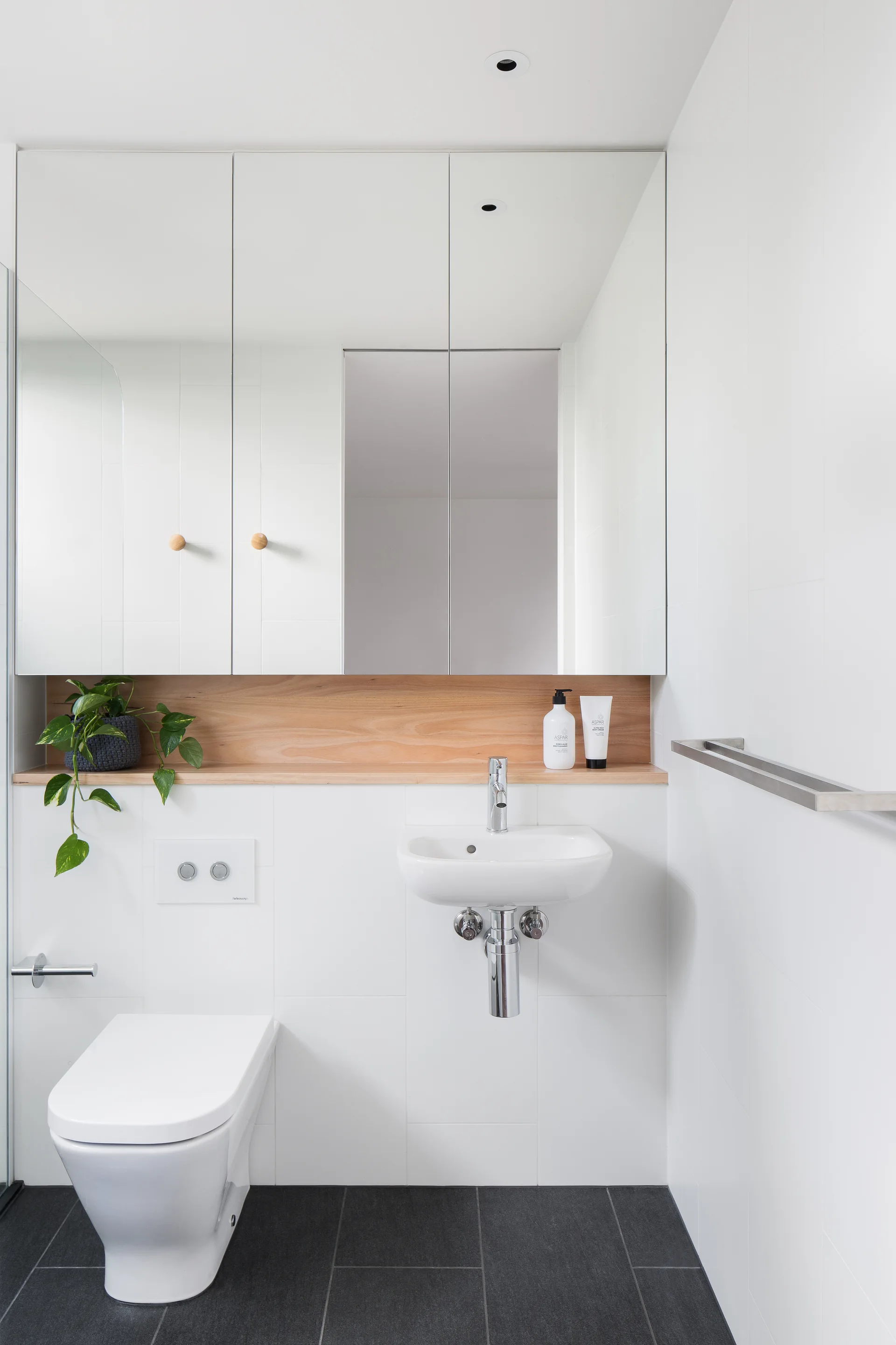 A minimalist bathroom with white tiled walls, a wall-hung toilet, a floating sink, and a mirrored medicine cabinet above a wood niche shelf.
