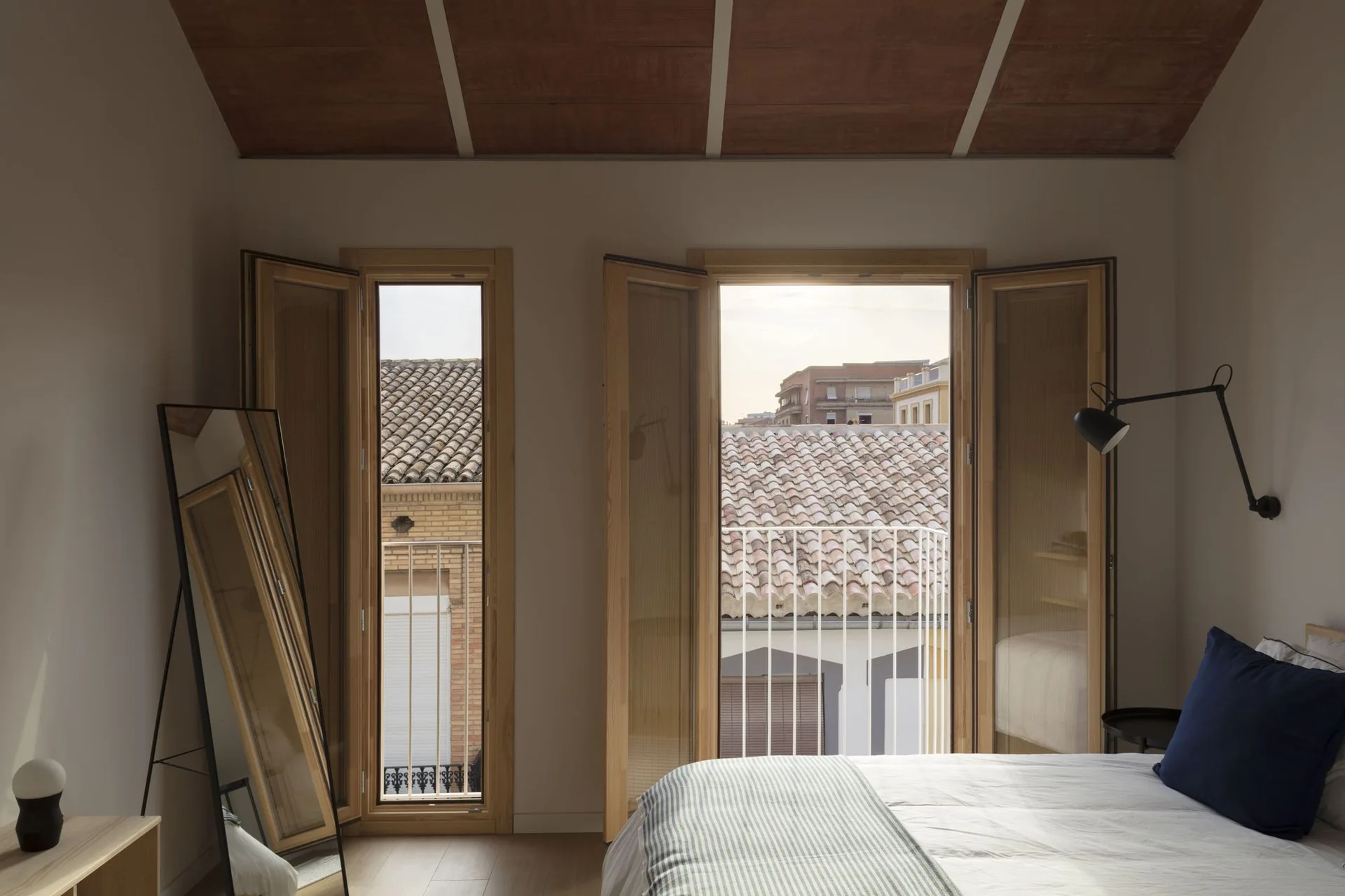 A minimalist bedroom featuring open wooden casement windows overlooking tile roofs, a bed with a blue pillow, a leaning floor mirror, and a wood ceiling.