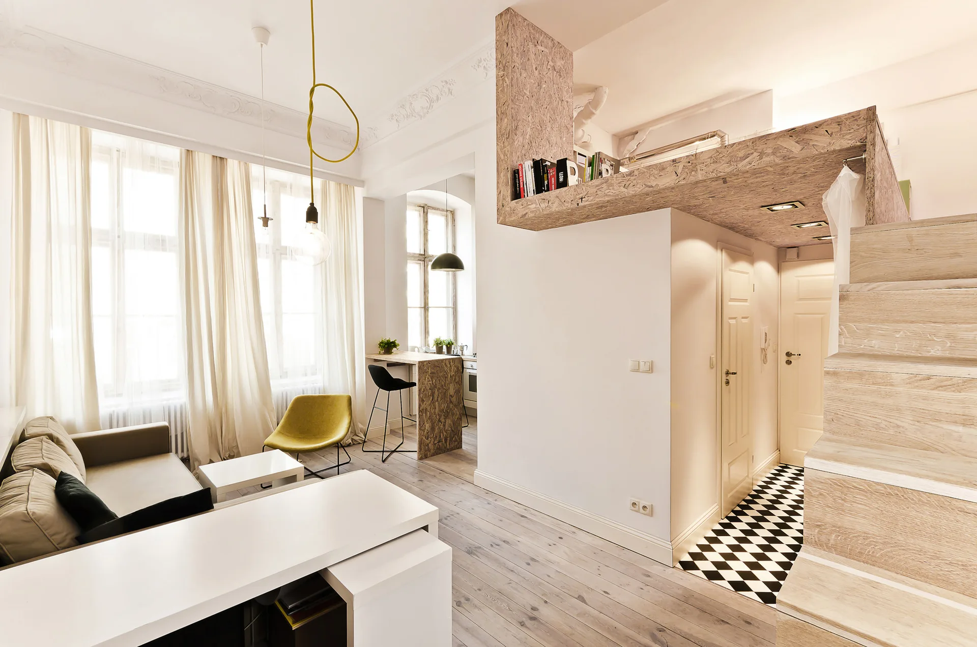 A compact studio apartment features a raw OSB loft platform, a light wood staircase, a small living area, and a checkerboard entry floor.