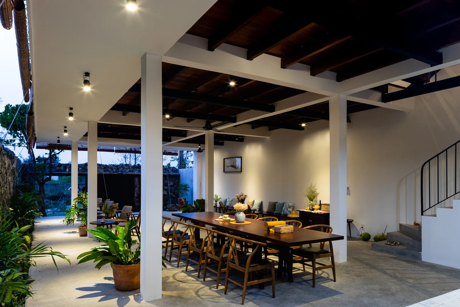 A long wooden dining table and chairs sit on a concrete patio under a dark wood beamed ceiling supported by white columns.