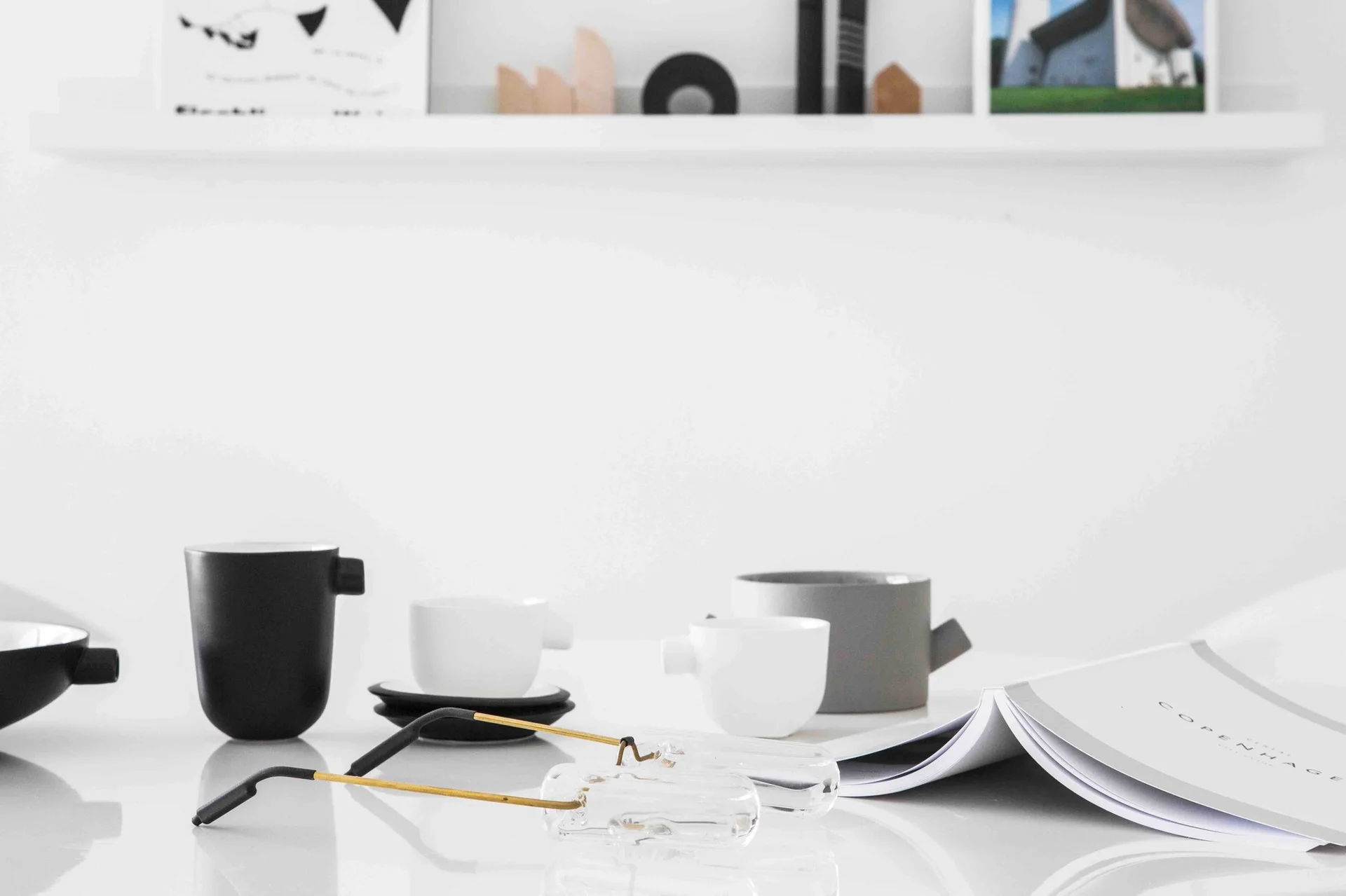 Matte black, grey, and white ceramic mugs rest on a white tabletop next to clear eyeglasses and an open book.