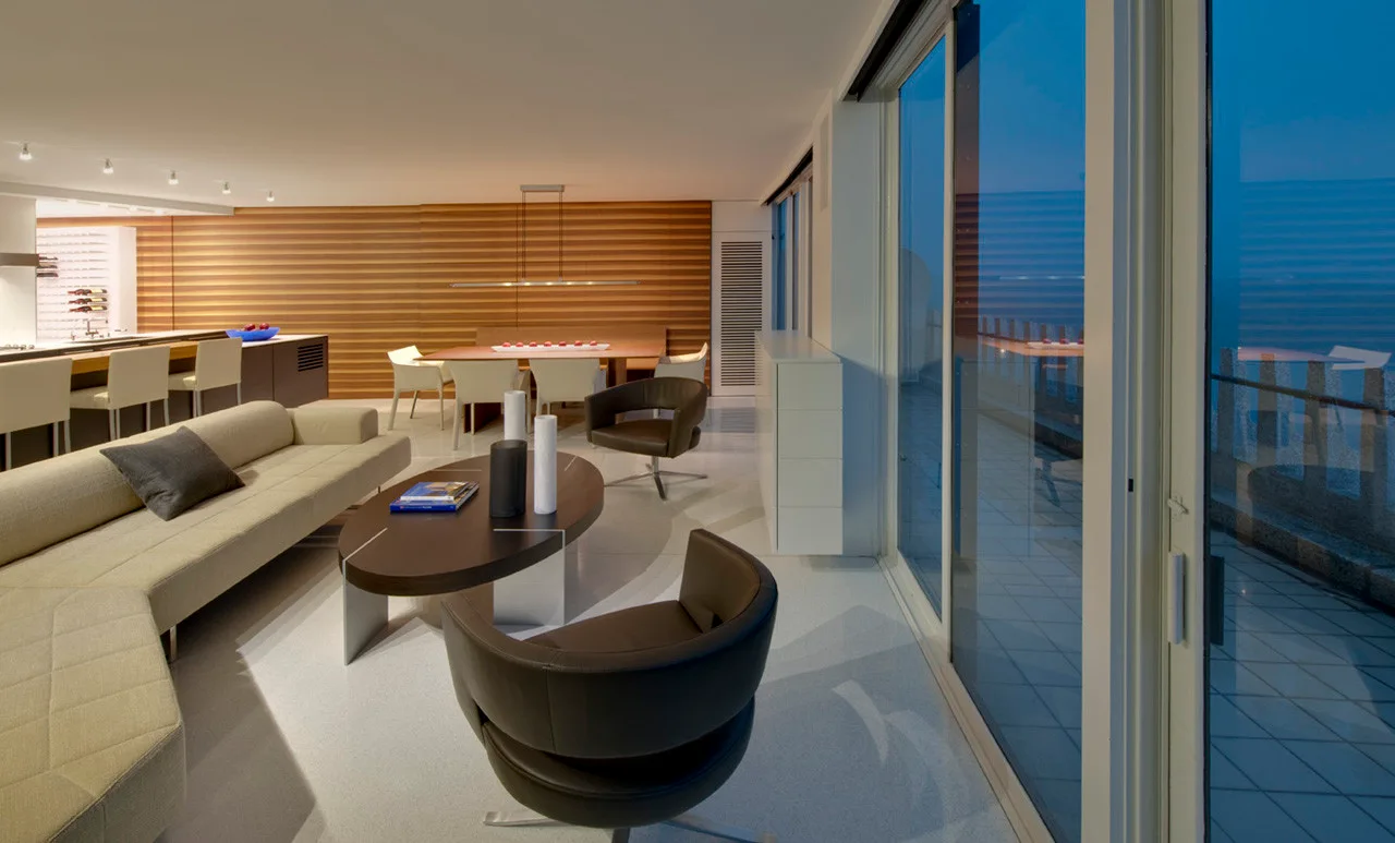 A modern open-concept living room features a low sectional sofa, dark swivel armchairs, and a warm wood slat wall beside sliding glass balcony doors.