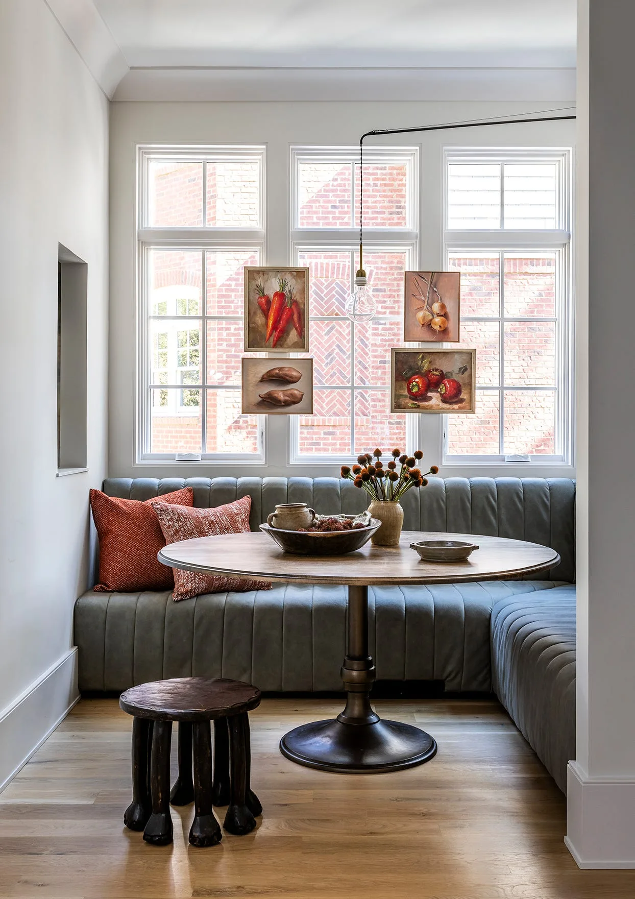 A round wooden pedestal table sits in a grey channel-tufted banquette nook beneath windows displaying four small framed paintings.