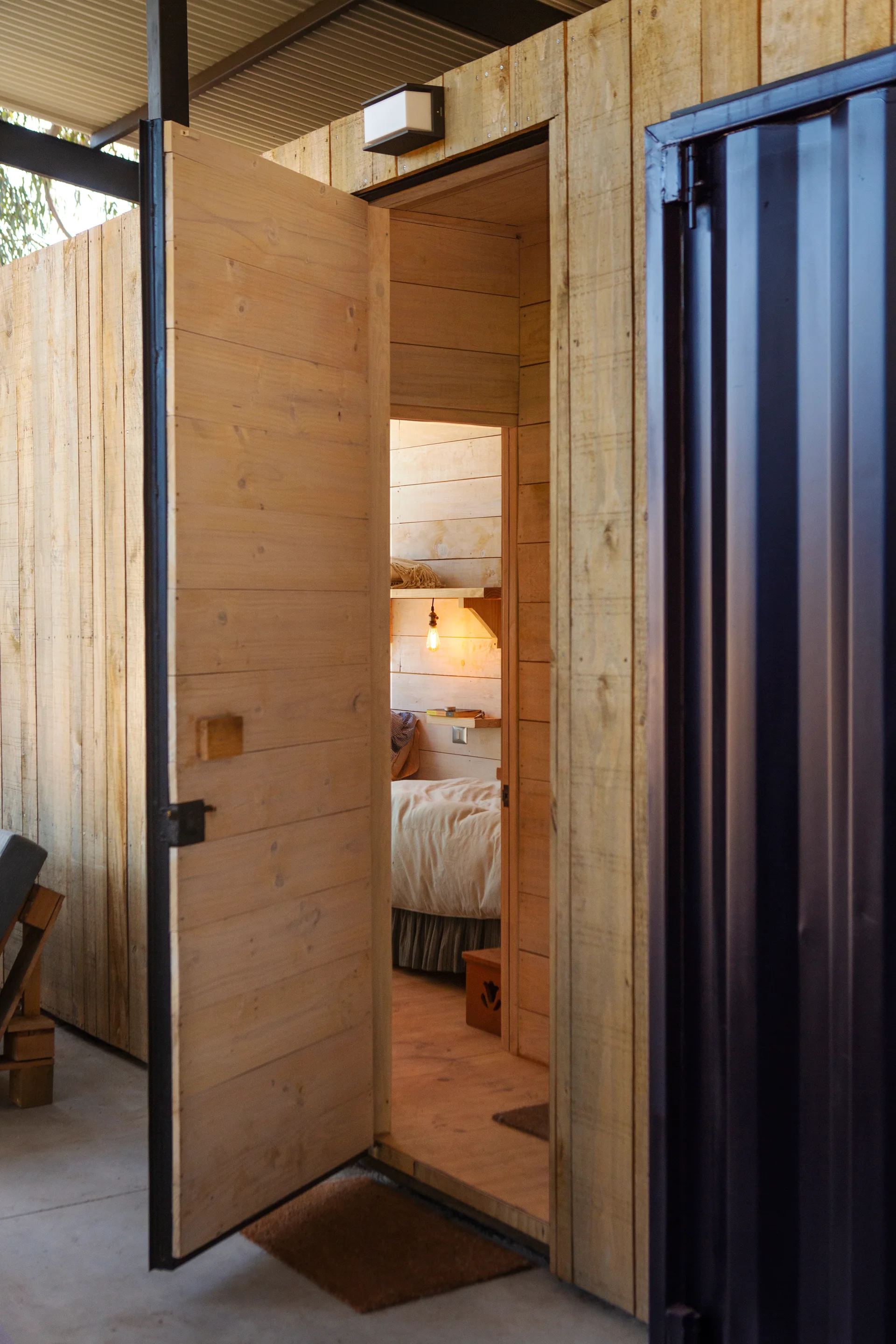 An open wood door reveals a small cabin bedroom featuring horizontal wood plank walls, a bed with linens, and a glowing hanging light bulb.