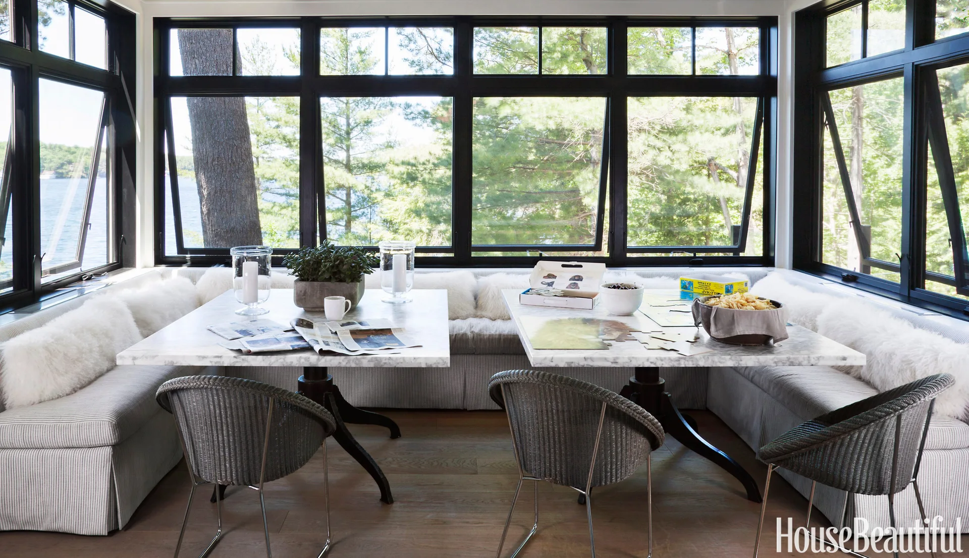 A sunlit dining nook features a u-shaped striped banquette, two marble pedestal tables, wicker chairs, and large black-framed windows overlooking trees.