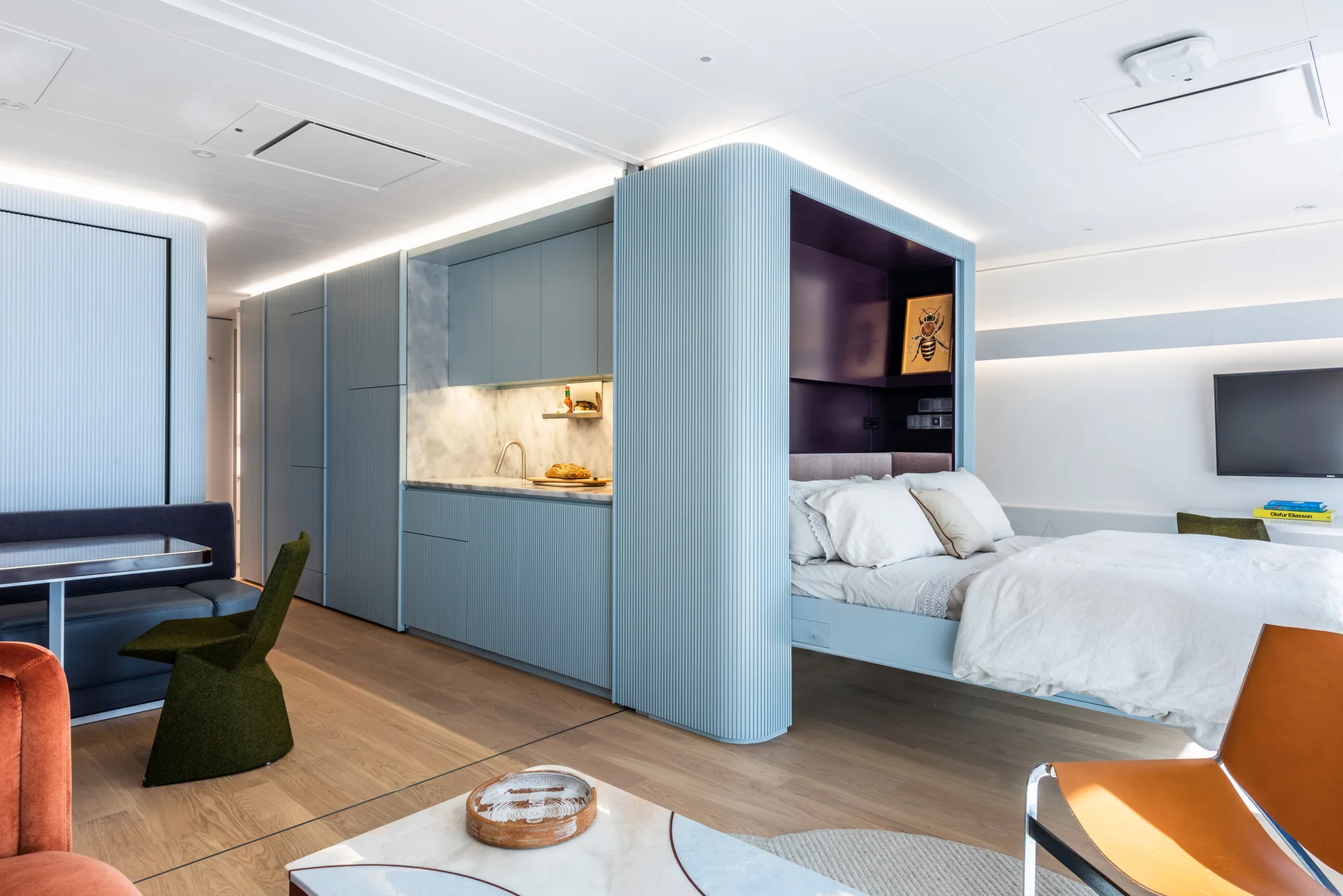 A modern studio apartment featuring a light blue fluted partition wall enclosing a bed niche next to an integrated kitchen.