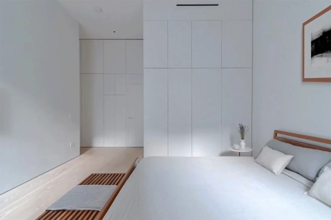 A minimalist bedroom features integrated white floor-to-ceiling cabinetry, a wooden bed, and a slatted timber bench at the foot of the bed.