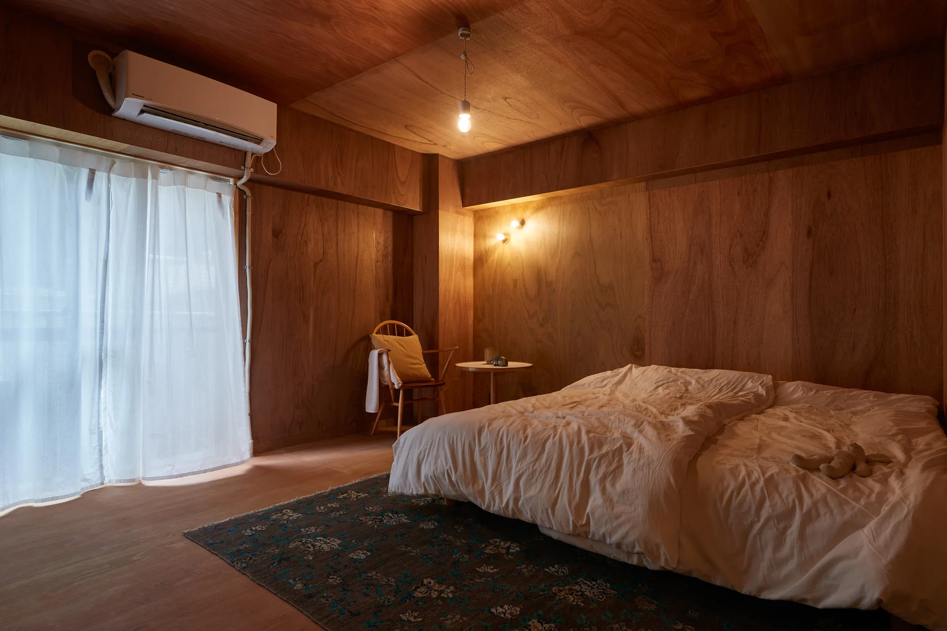 A minimalist bedroom features walls and ceilings covered in warm wood paneling, a low bed with white linens, and a dark patterned rug.