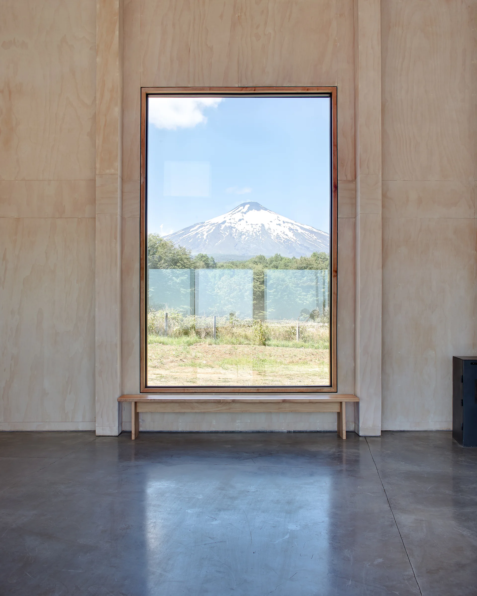 A minimalist room featuring light plywood paneled walls, a polished concrete floor, and a large picture window framing a snow-capped mountain.