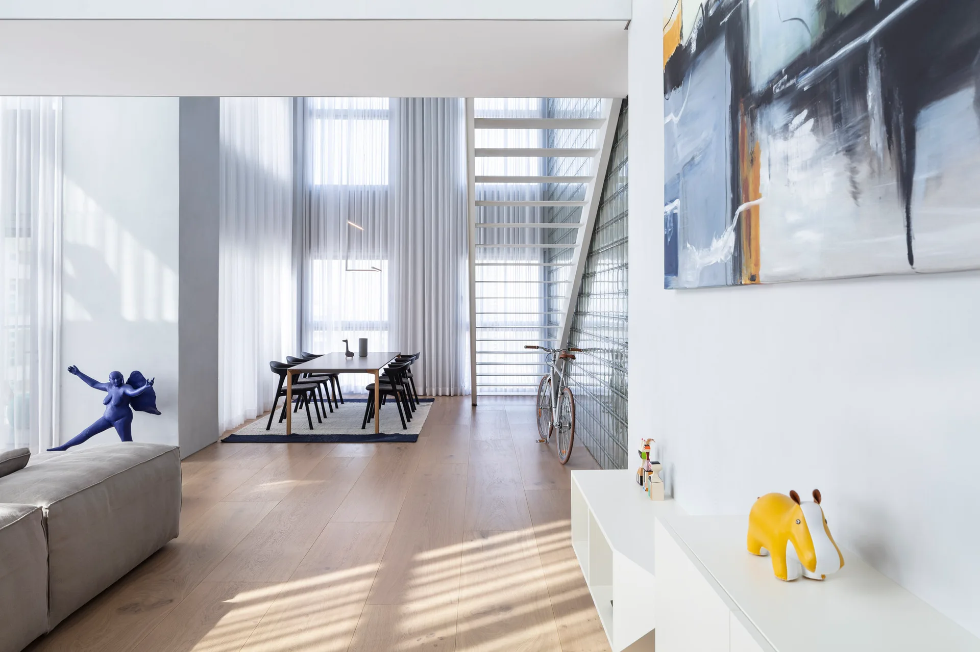 A bright dining area features a wooden table and black chairs next to a white open staircase and a translucent glass block wall.