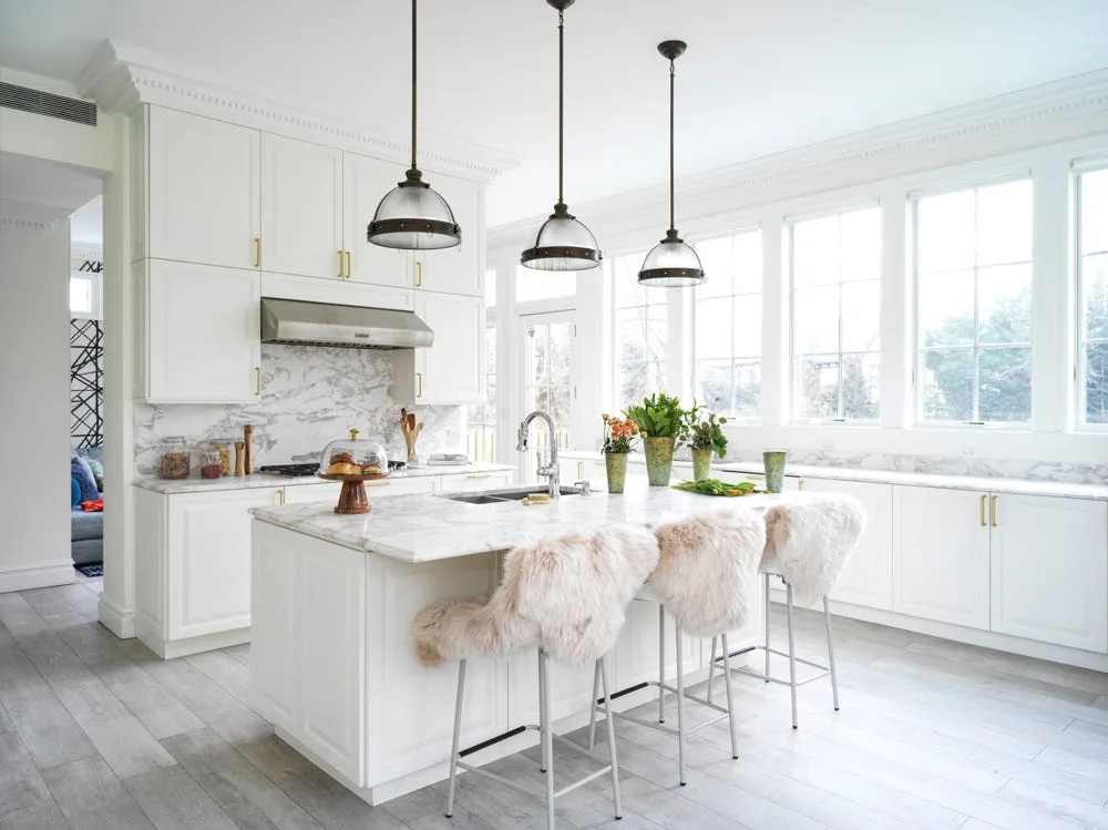 A bright white kitchen featuring a marble-topped island, three barstools with fluffy white covers, and three dome pendant lights.