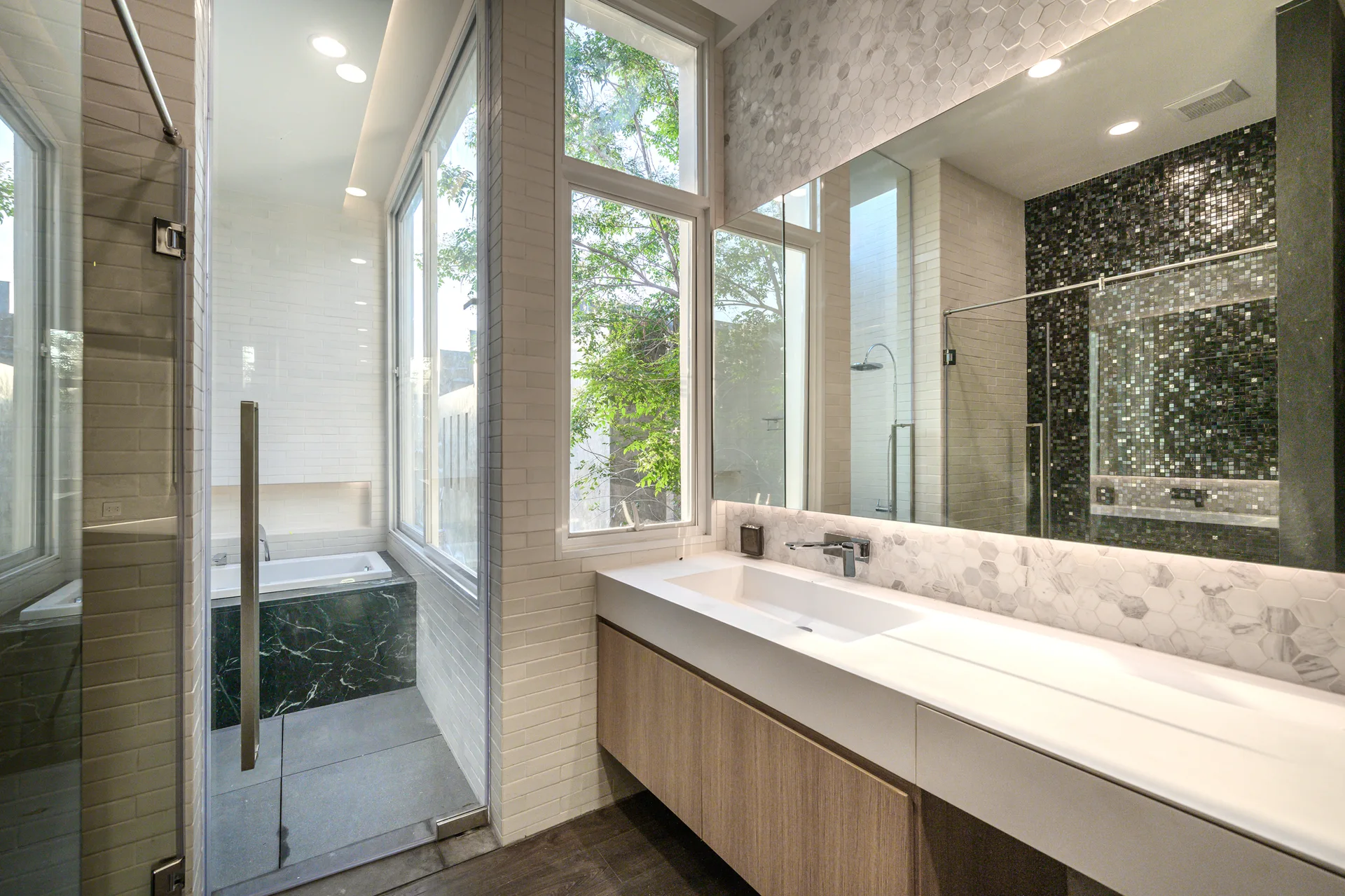 A modern bathroom showcases a floating wooden vanity, a large frameless mirror with hexagonal tile backsplash, and a glass-enclosed bathtub area.