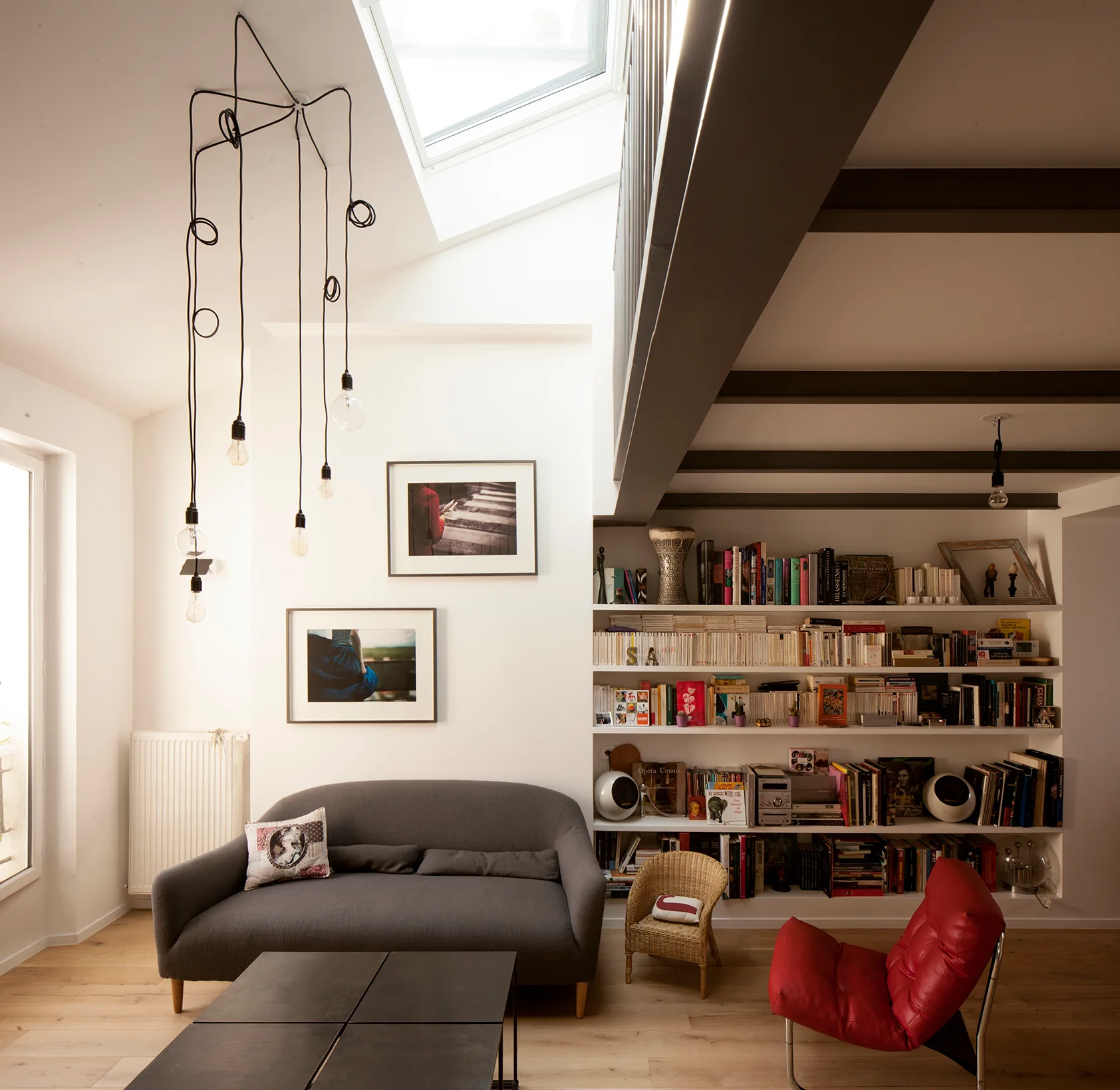 A bright living room features a grey sofa, a red lounge chair, white built-in bookshelves, and a large skylight next to dark ceiling beams.