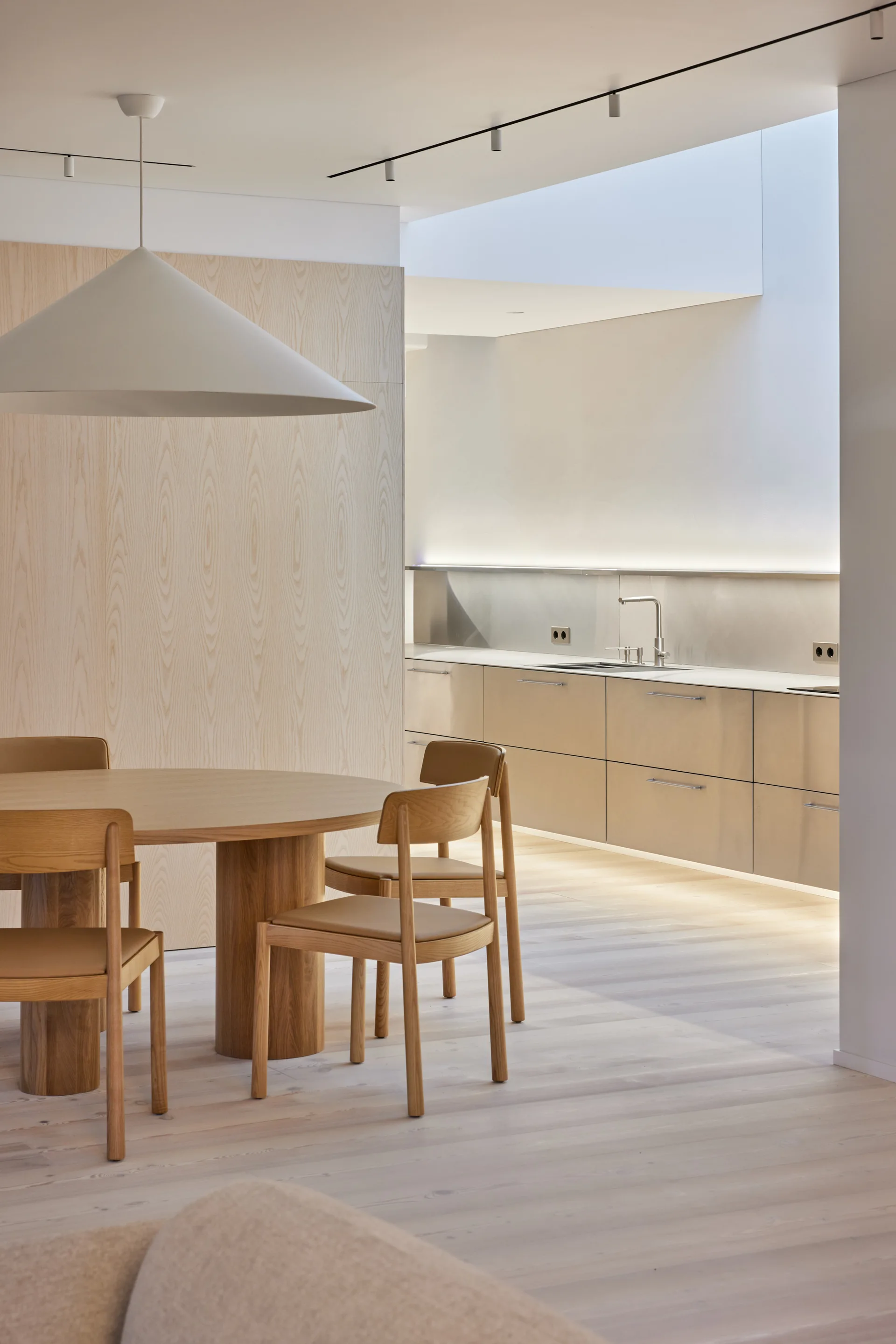 A modern round dining table and matching chairs sit before a light-wood paneled wall and sleek metal kitchen cabinetry under a skylight.