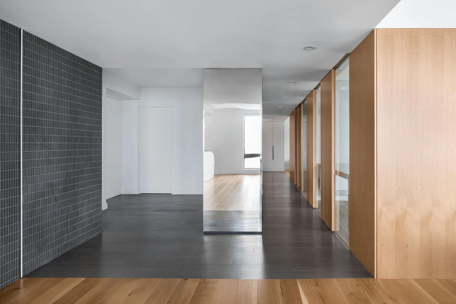 A minimalist hallway featuring a central floor-to-ceiling mirrored column, a dark grey tiled feature wall, and light wood paneled cabinets.
