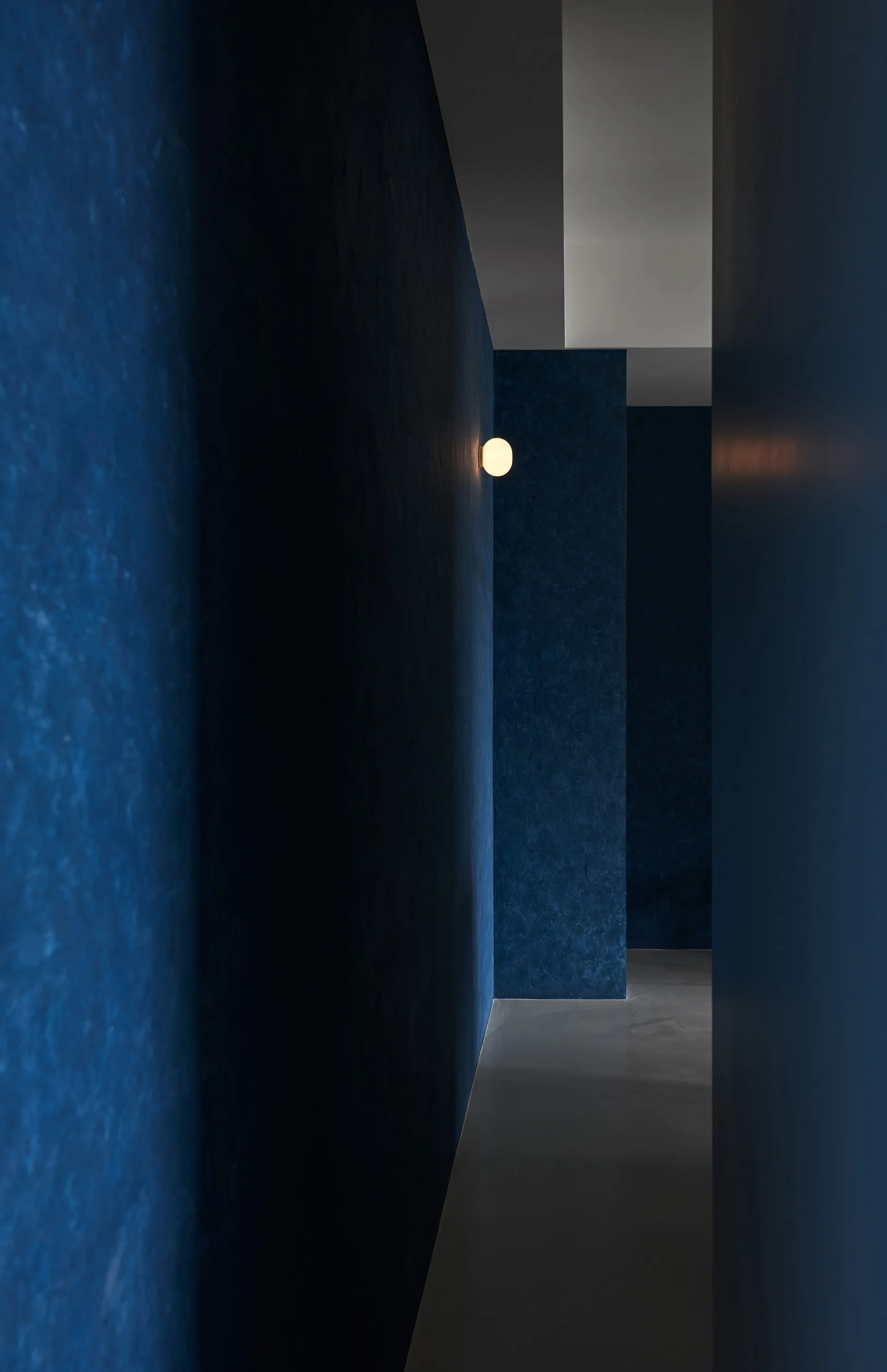 A narrow hallway features mottled blue plaster walls, a smooth grey floor, and a single glowing white globe sconce.