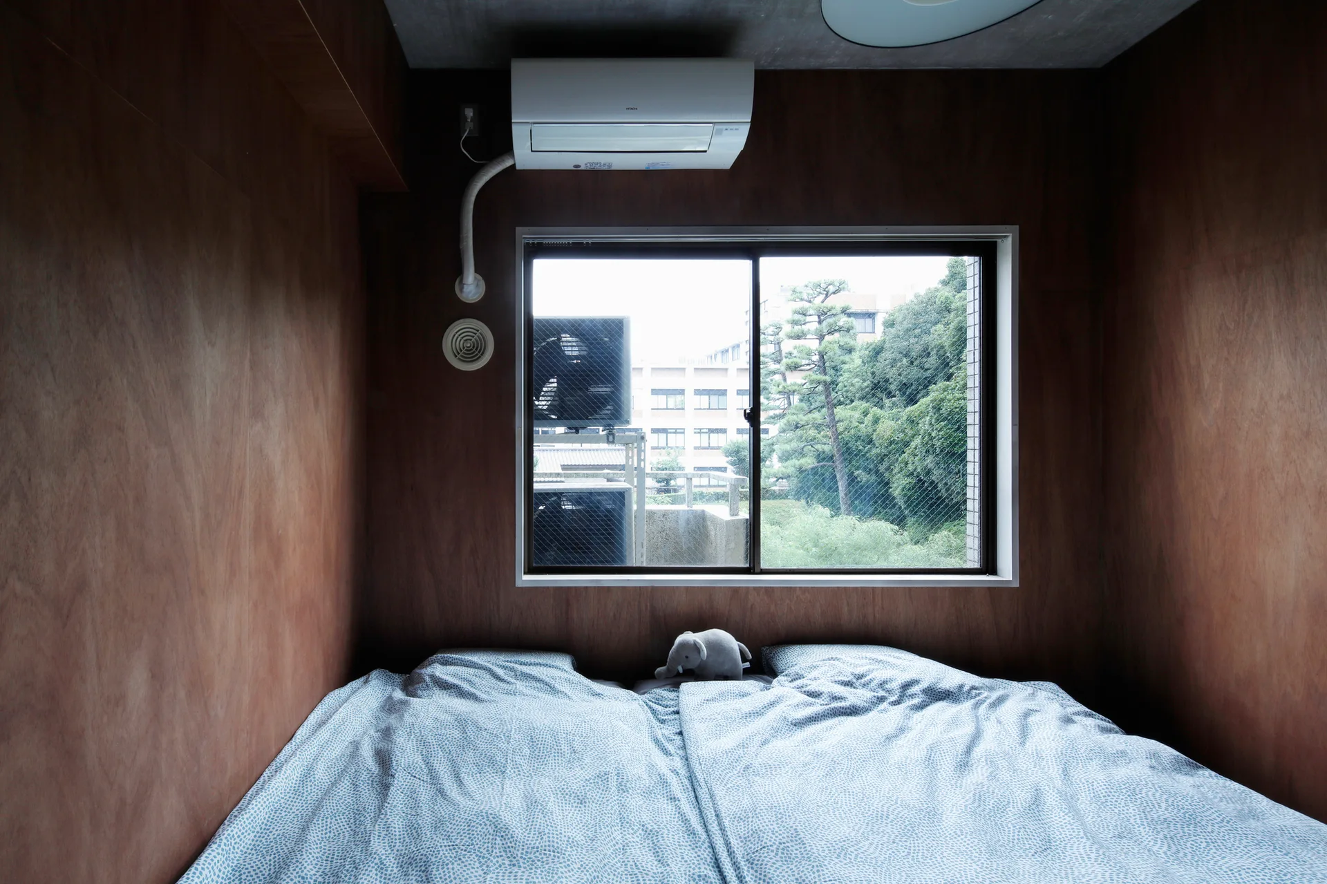 A compact bedroom features dark wood-paneled walls, a gray concrete ceiling, and a large bed with blue patterned sheets under a window.
