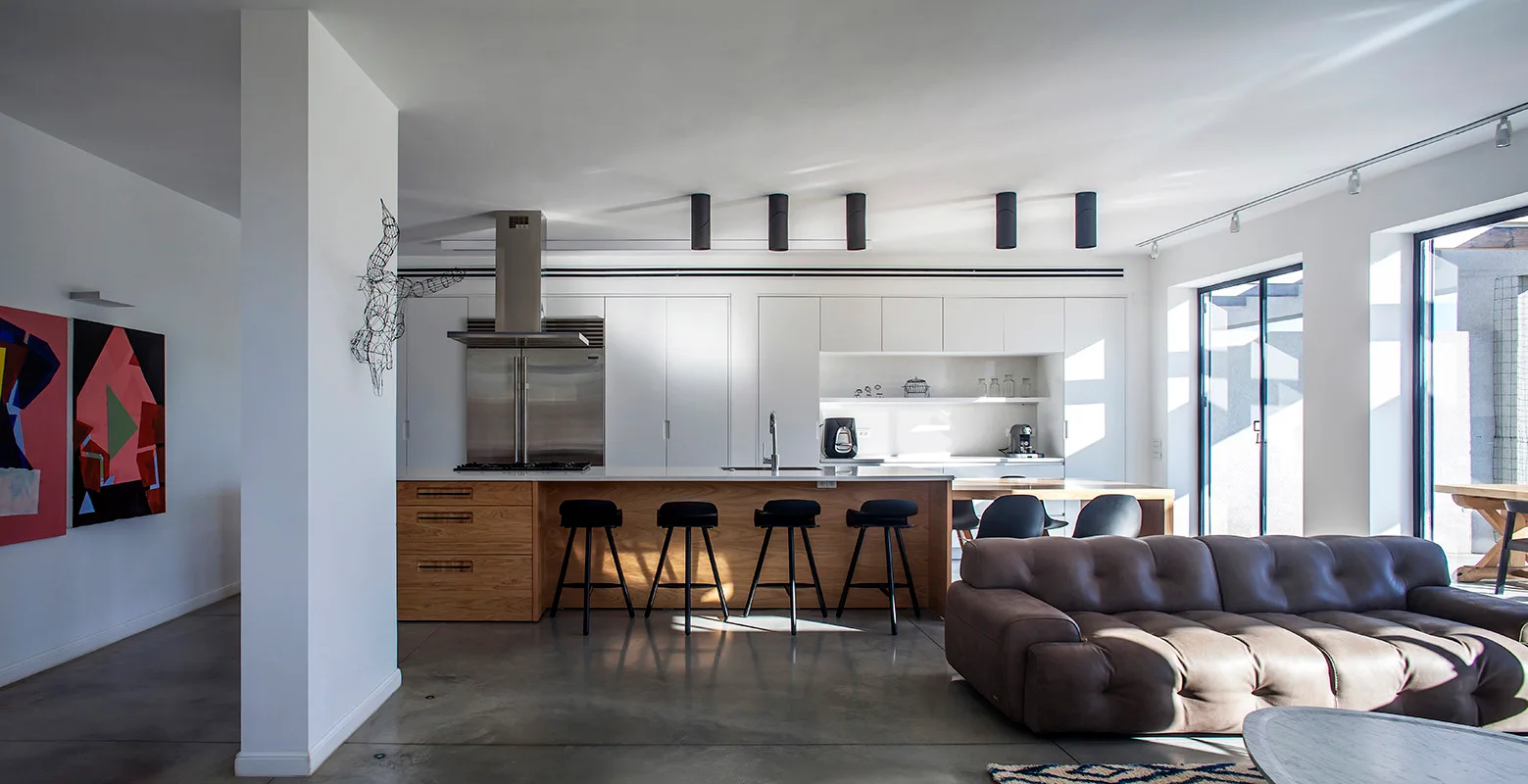 A modern open-plan kitchen and living space featuring a wood kitchen island with black stools, seamless white cabinets, and a brown tufted sofa.