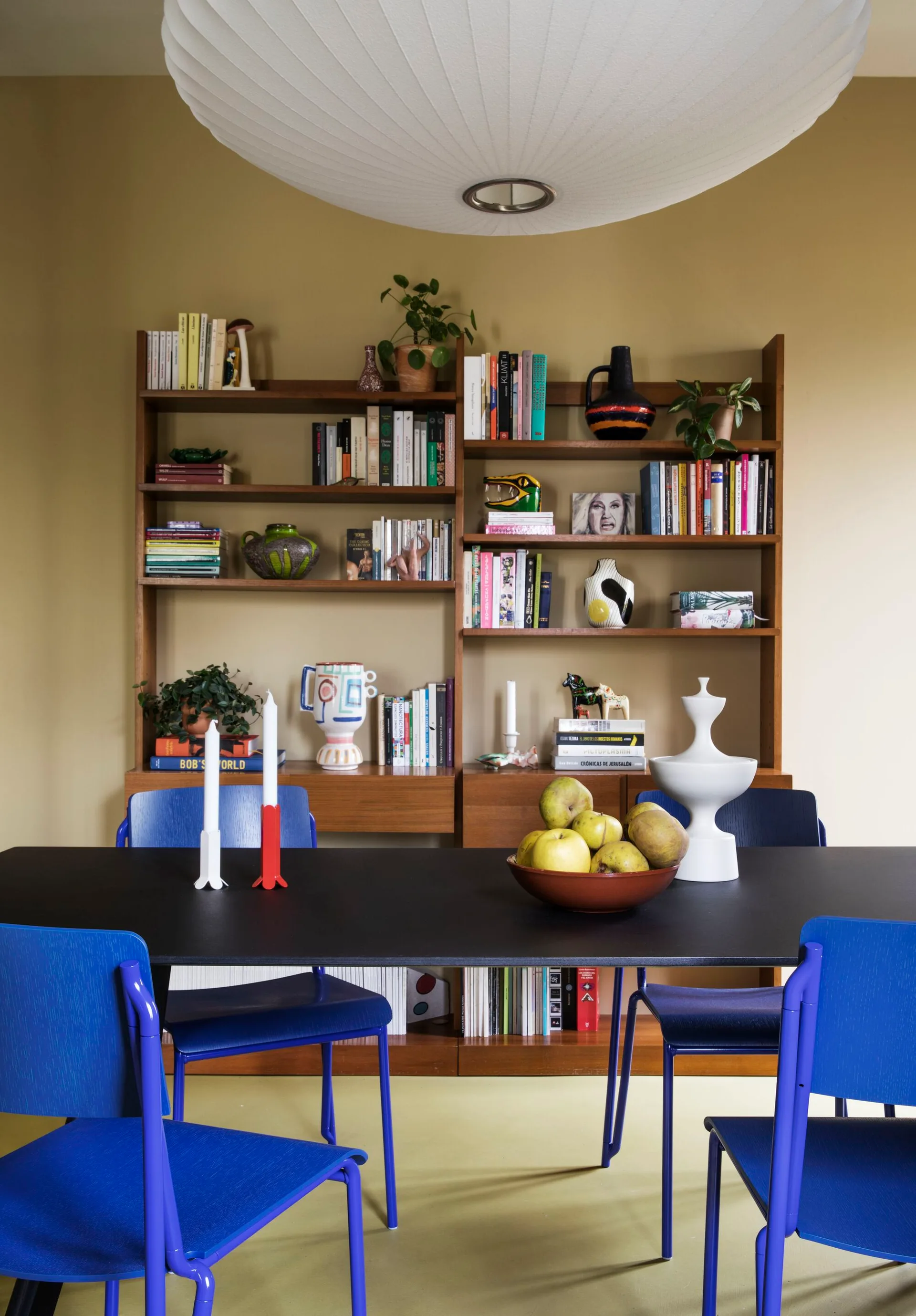 Vibrant blue dining chairs surround a black dining table in front of a large wooden bookshelf against a mustard yellow wall.