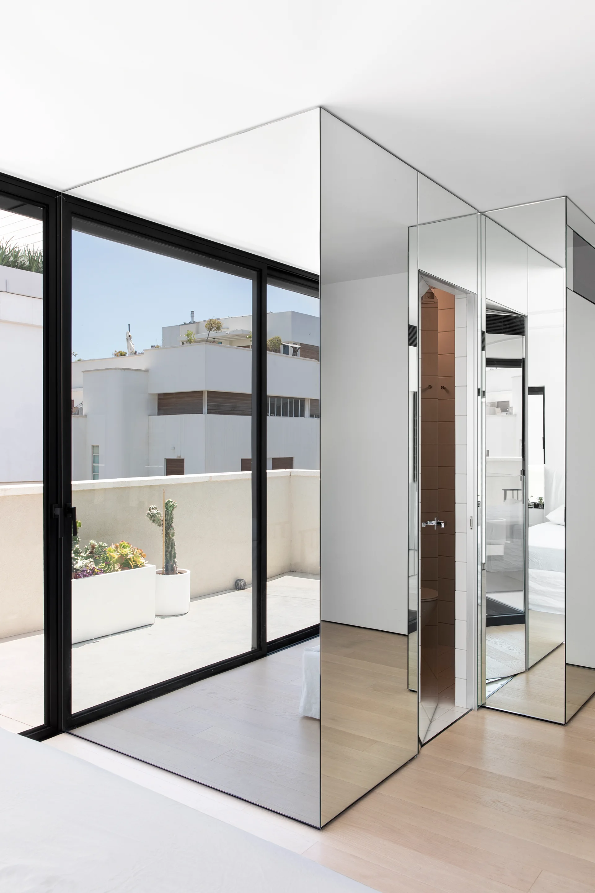 A minimalist bedroom featuring floor-to-ceiling mirrored walls that reflect light from black-framed sliding glass doors leading to a sunny terrace.