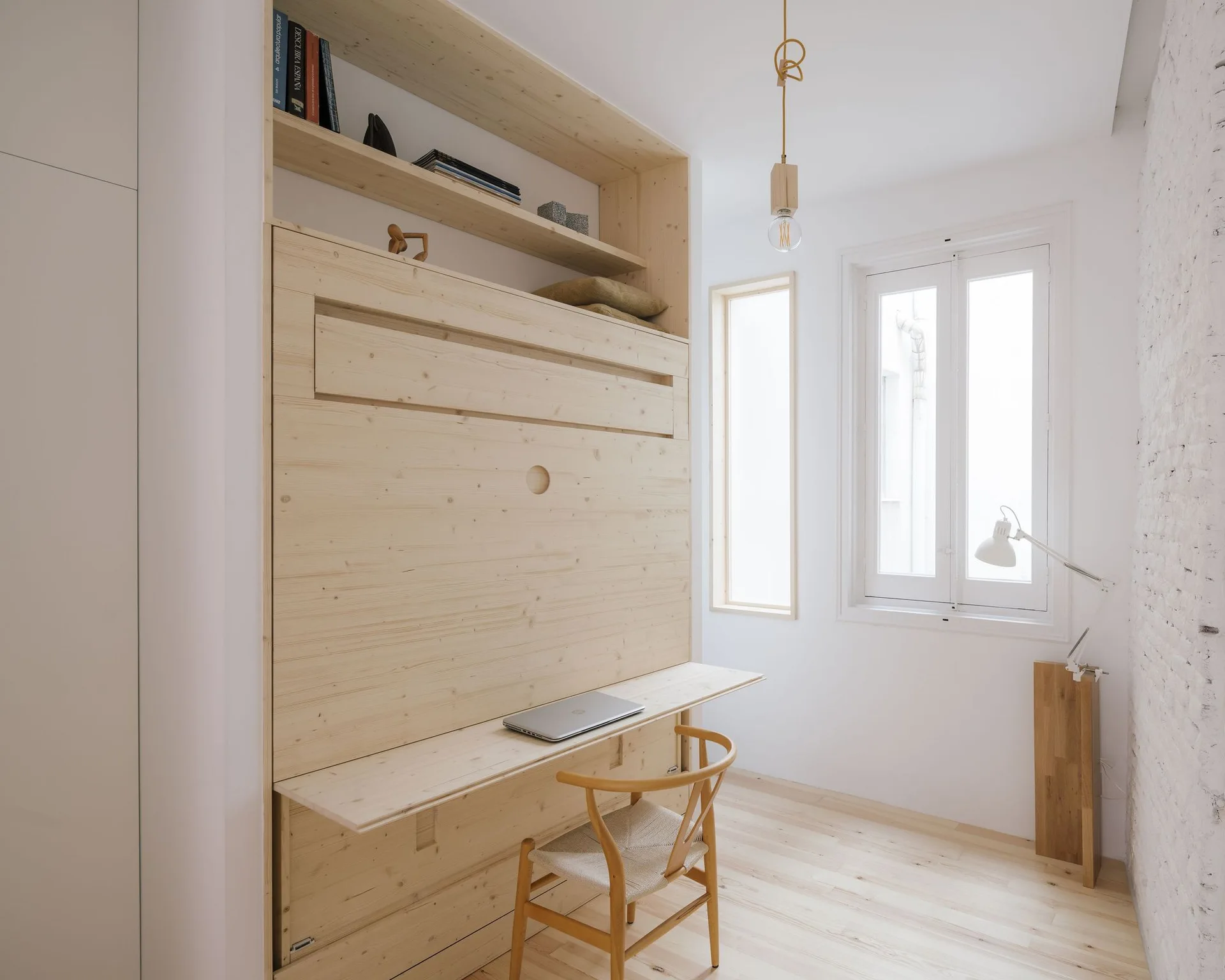 A light wood Murphy bed with an integrated fold-down desk and a matching wooden chair sits in a bright room next to a white brick wall.