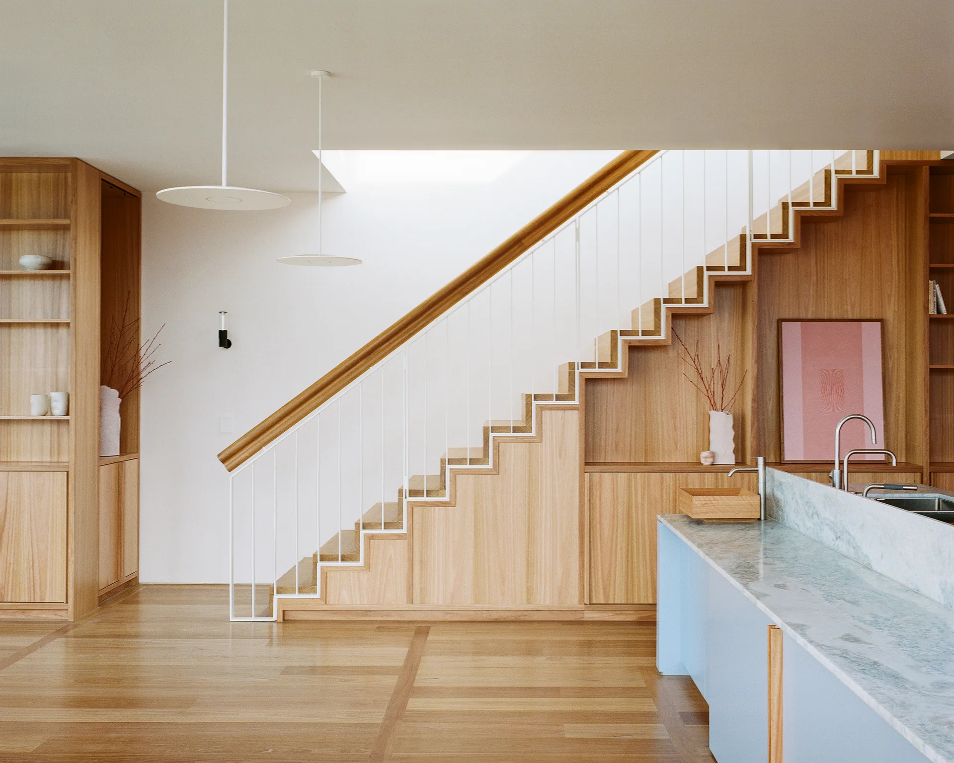 A light wood staircase with integrated storage cabinets sits next to a pale blue kitchen island under flat white pendant lights.