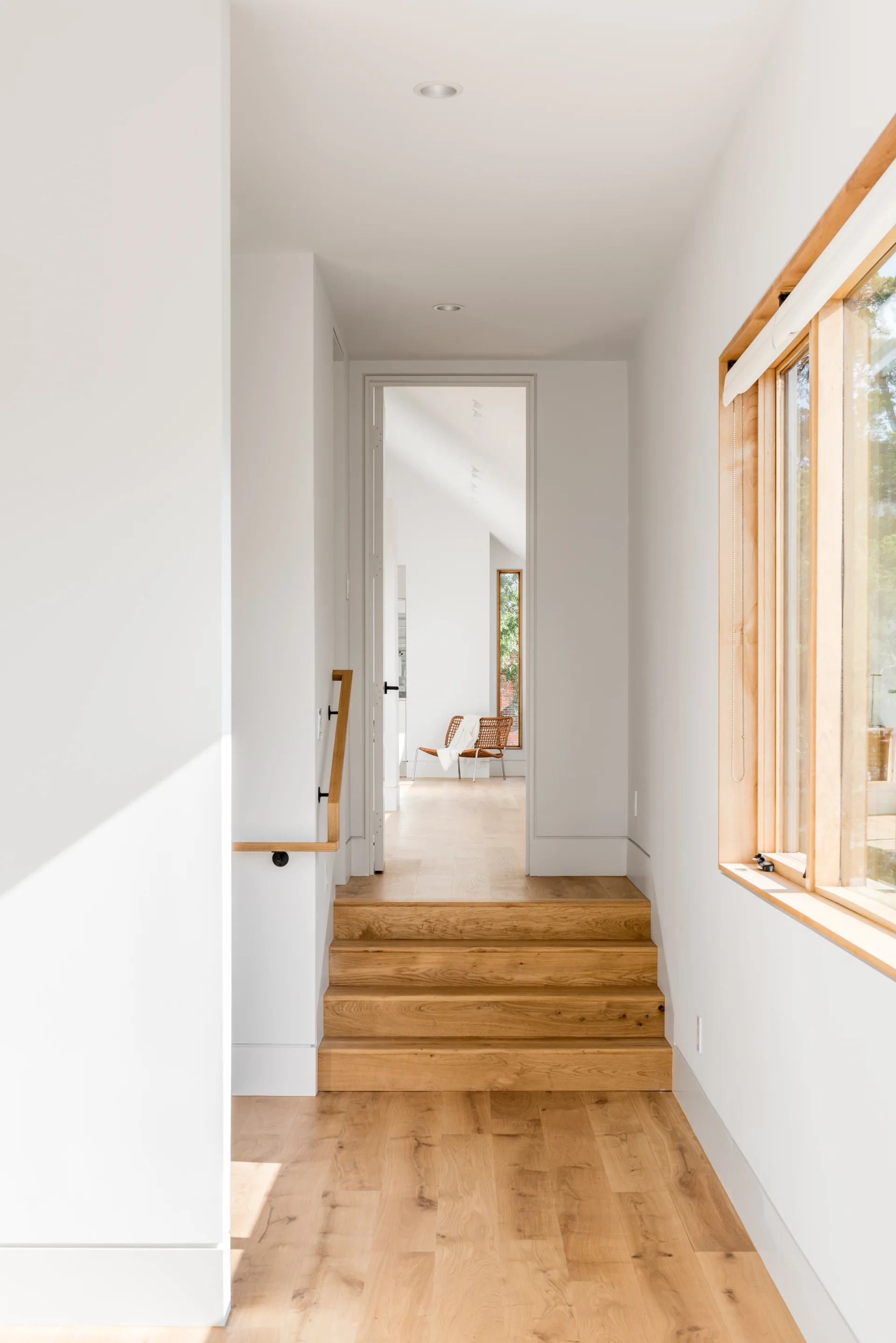 A minimalist white hallway features light wood stairs, matching hardwood floors, and a natural wood-framed window.