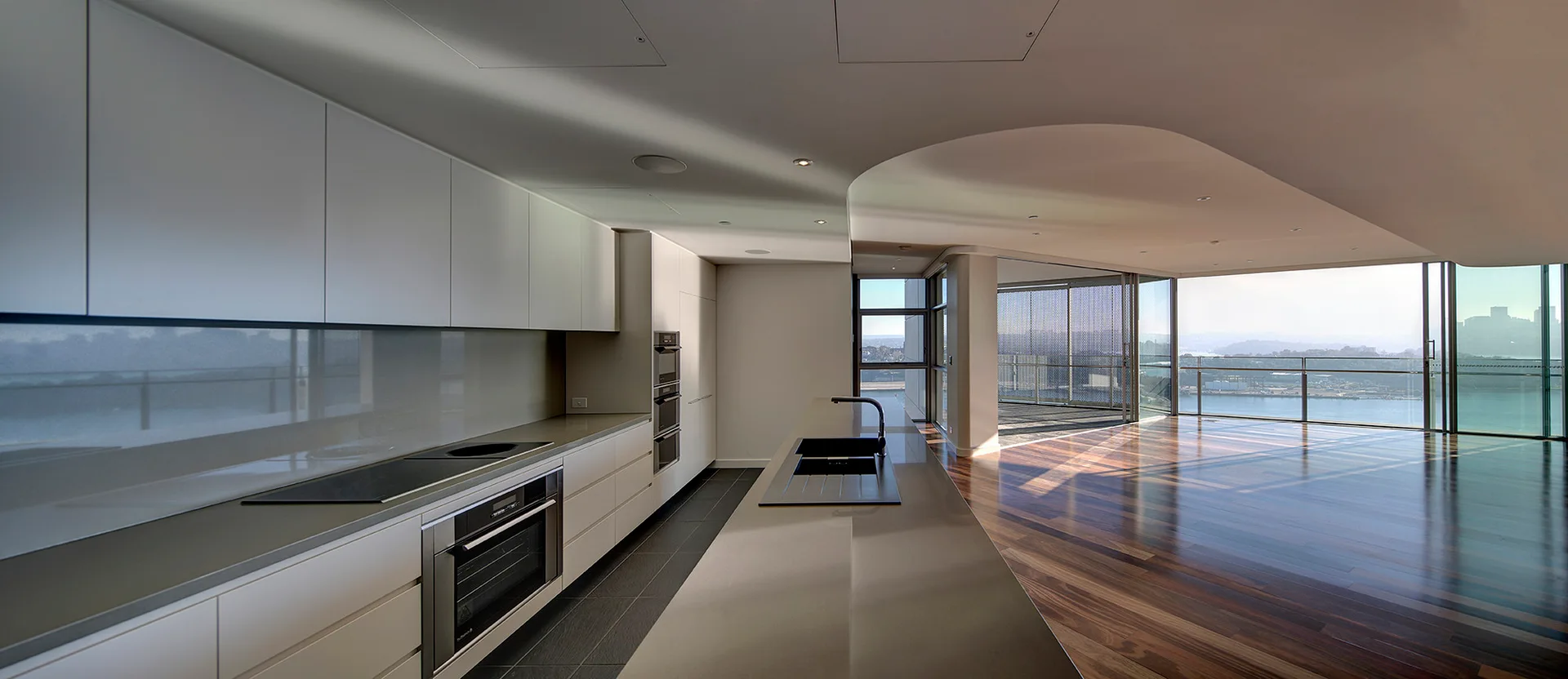 A minimalist kitchen featuring white flat-panel cabinetry, a long gray island with a sink, and floor-to-ceiling windows with a water view.