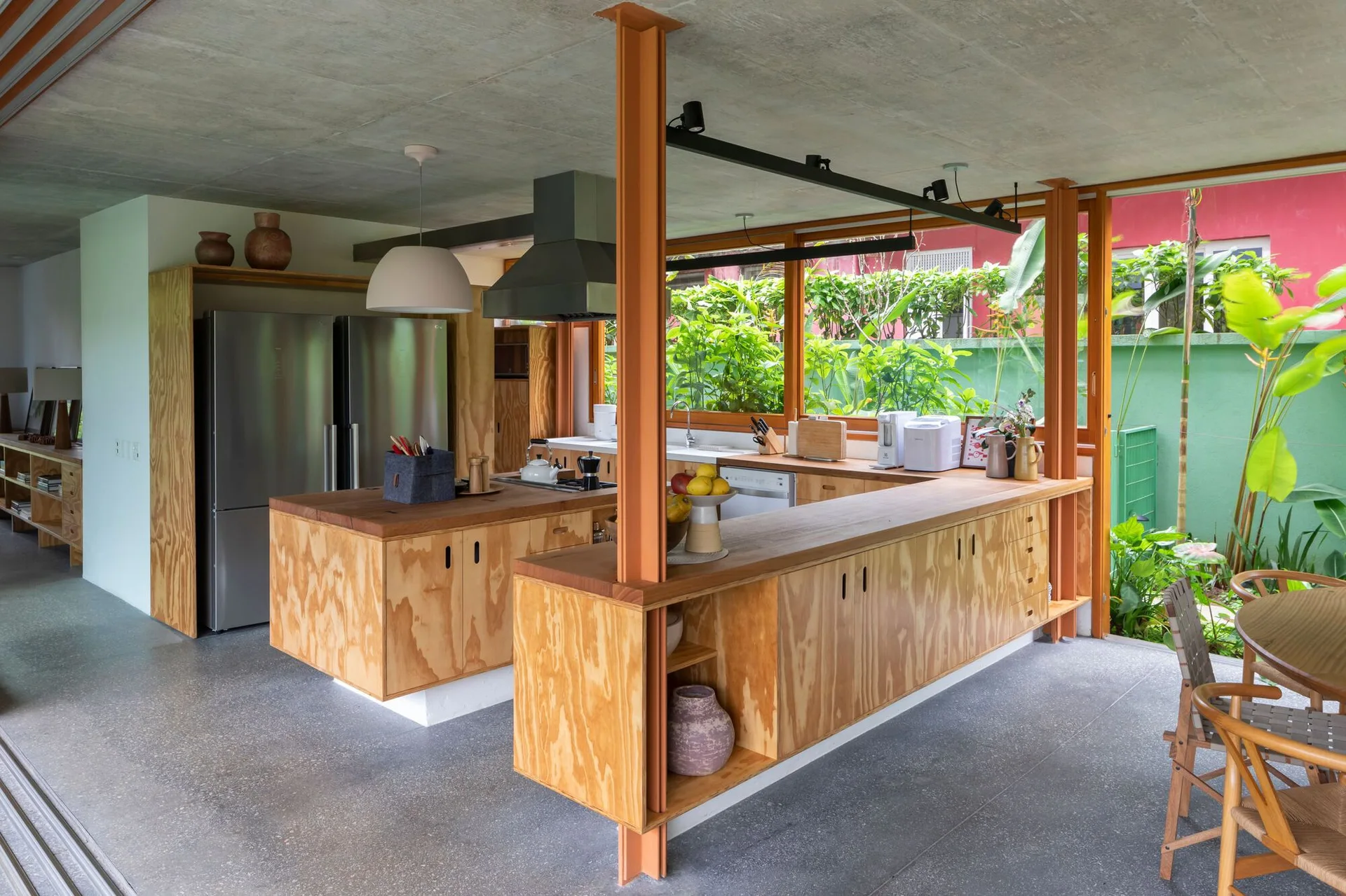 An open-plan kitchen featuring custom plywood cabinetry, a stainless steel refrigerator, orange steel support columns, and an exposed concrete ceiling.