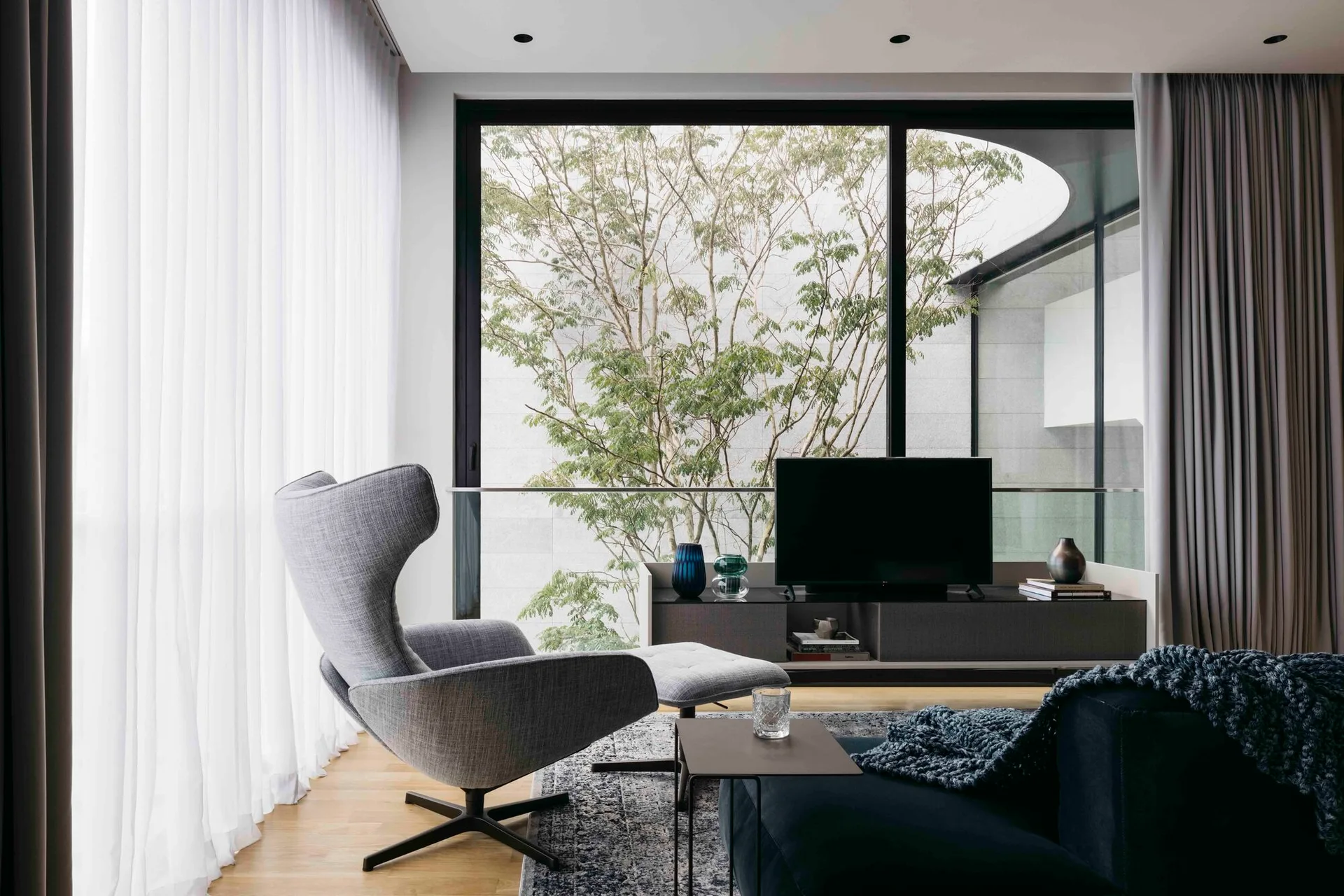 A modern grey wingback armchair and ottoman sit near a low media console in a living room with floor-to-ceiling windows showing a tree.