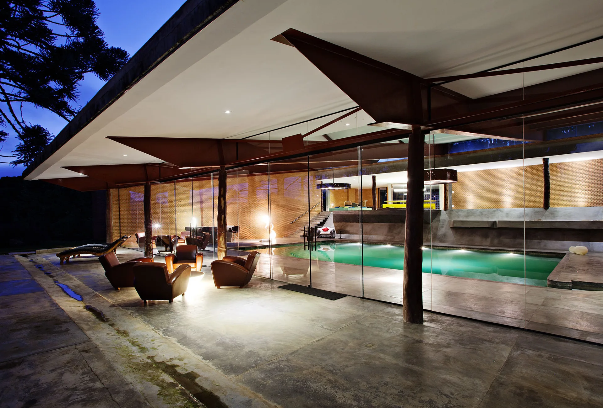 A covered concrete patio features brown leather armchairs separated from an indoor swimming pool by a large floor-to-ceiling glass wall.