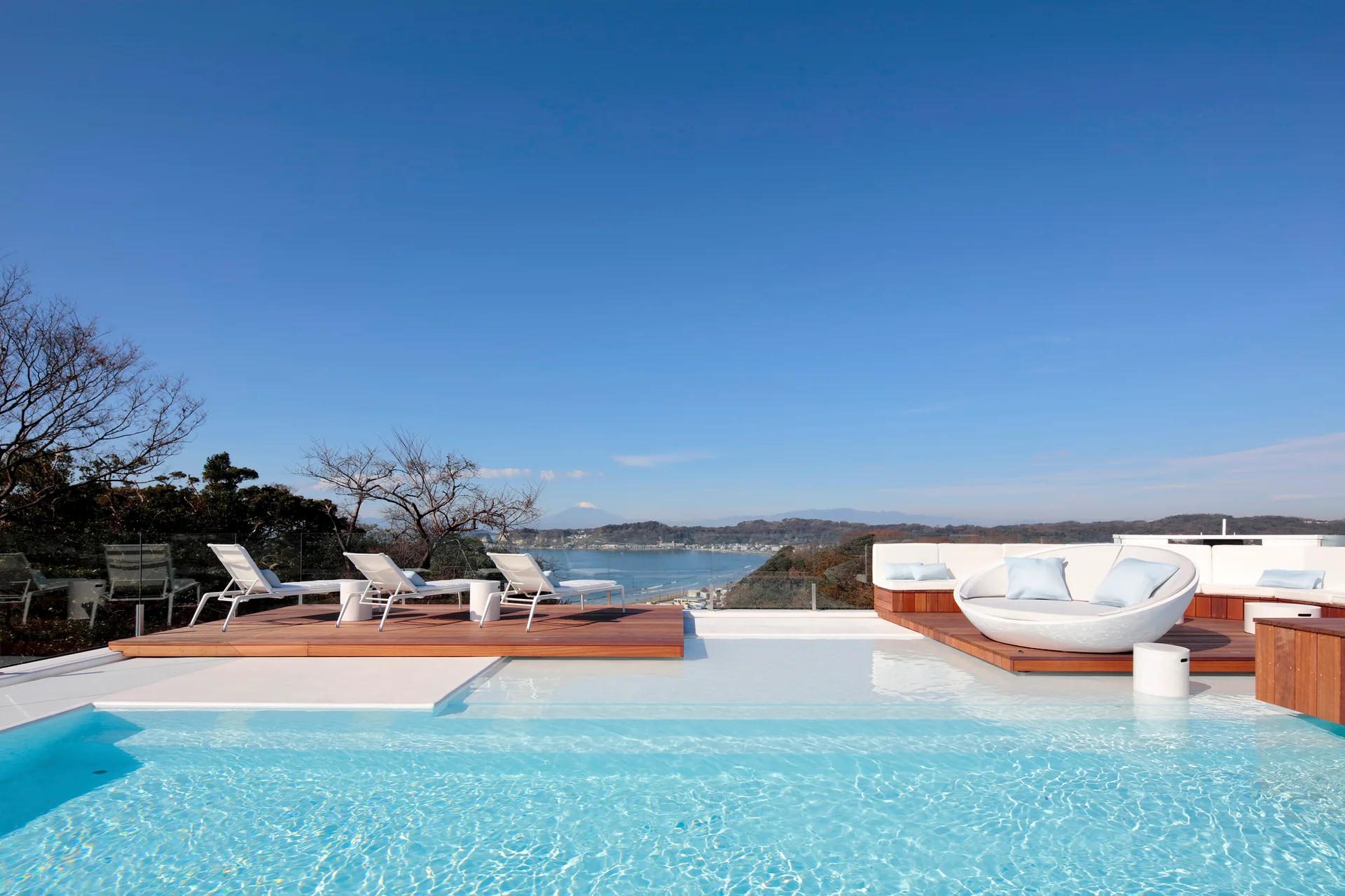 A rectangular turquoise swimming pool features a wooden deck with white sun loungers and a circular daybed overlooking a coastal landscape.