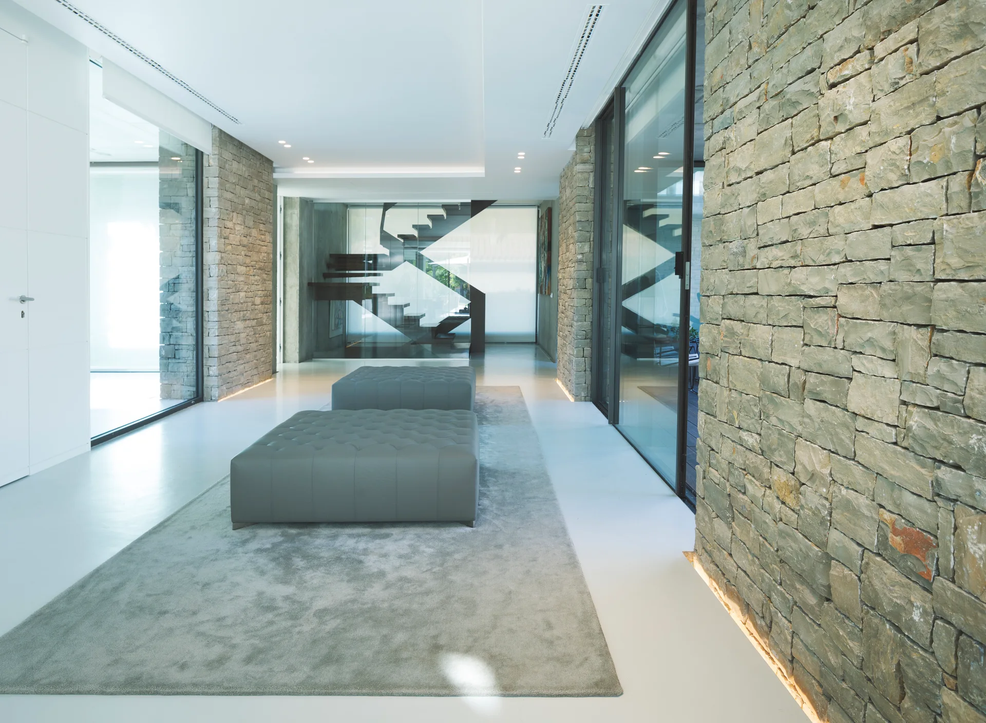 A spacious white hallway features two gray tufted ottomans on a rug between textured stone walls, with a modern staircase visible in the background.