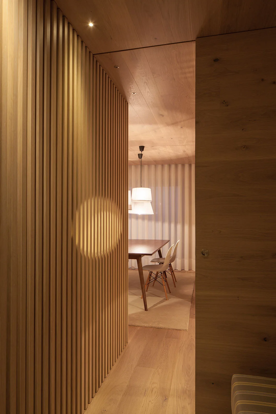 A modern dining room with a wooden table and chairs viewed past a vertical wood slat wall, featuring warm recessed lighting and a white pendant lamp.