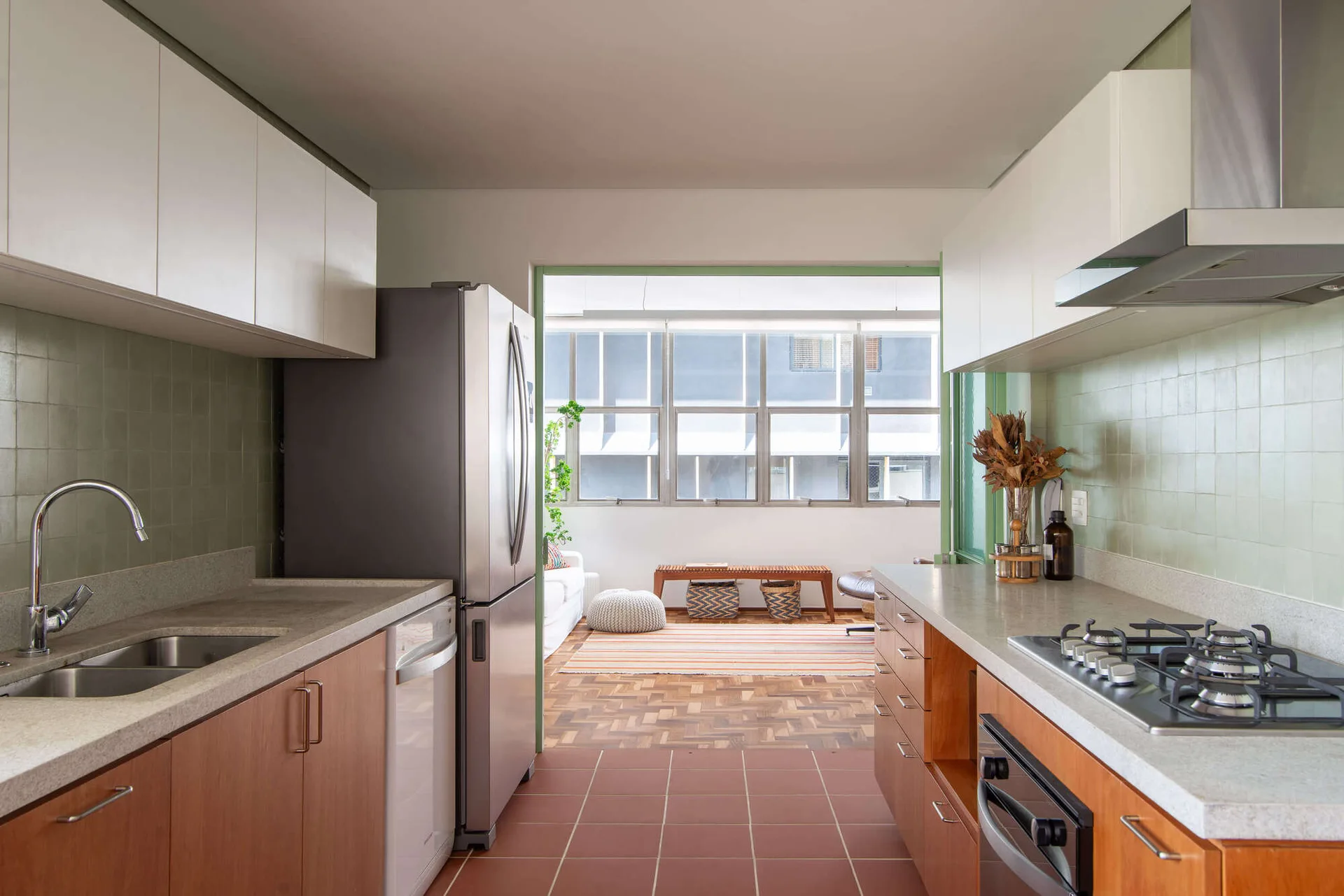 A galley kitchen features wood lower cabinets, white upper cabinets, and sage green tiled walls, looking into a sunlit living room with parquet flooring.