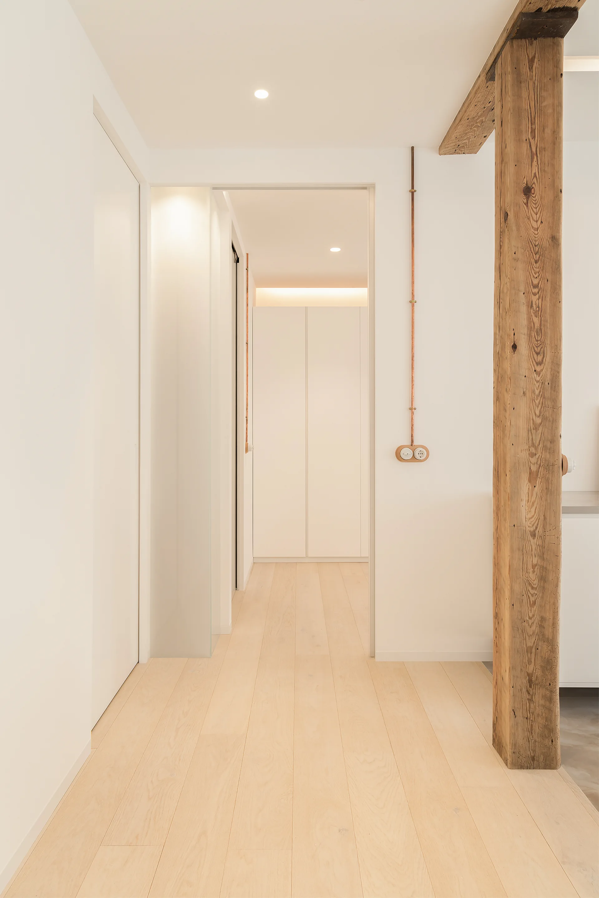 A minimalist hallway featuring light wood flooring, a rustic wooden support pillar, and vertical copper conduits on white walls.