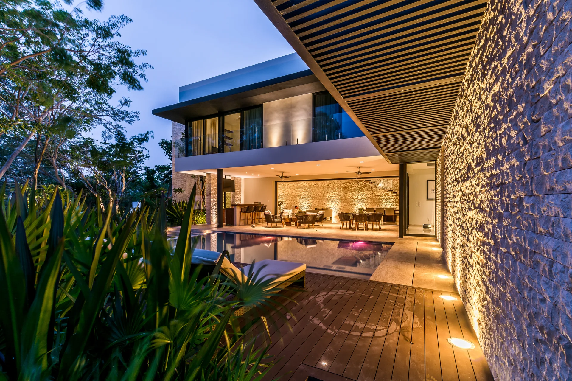 A modern patio at twilight featuring a swimming pool, lounge chairs, a slatted ceiling, and an uplit stacked stone accent wall.