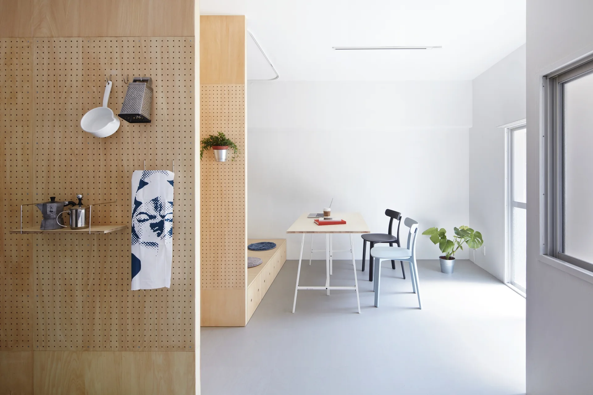 A minimalist dining area featuring a light wood table, two chairs, and a large plywood pegboard partition wall holding kitchen utensils.