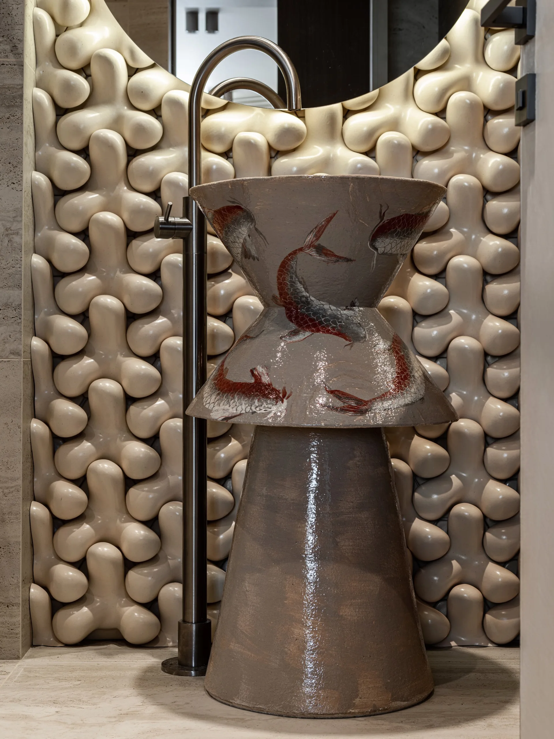A glazed ceramic hourglass pedestal sink featuring painted koi fish stands in front of a dimensional cream-colored textured tile wall.
