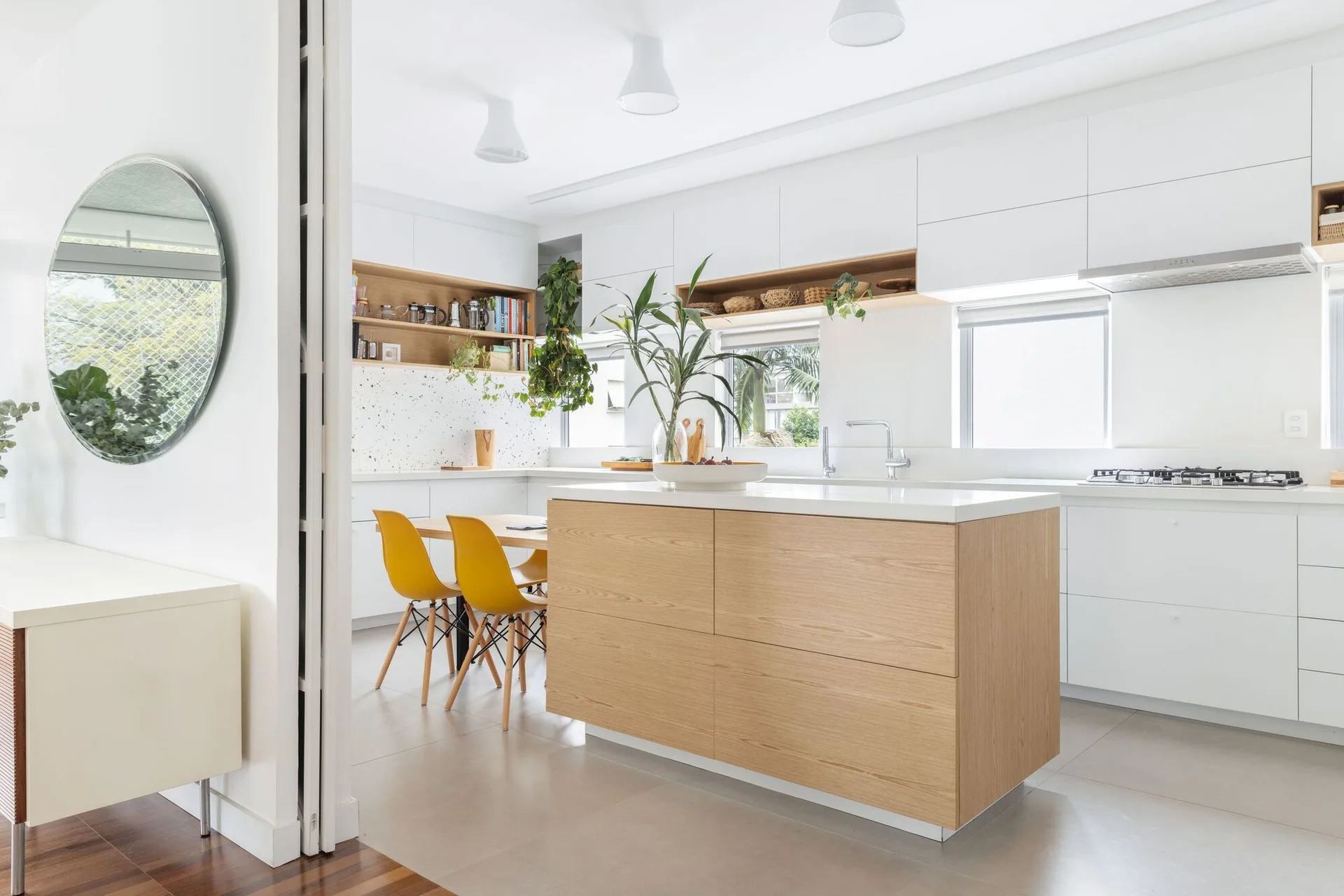 A bright modern kitchen features a wood-toned central island, white flat-panel cabinetry, a terrazzo backsplash, and a dining table with yellow chairs.