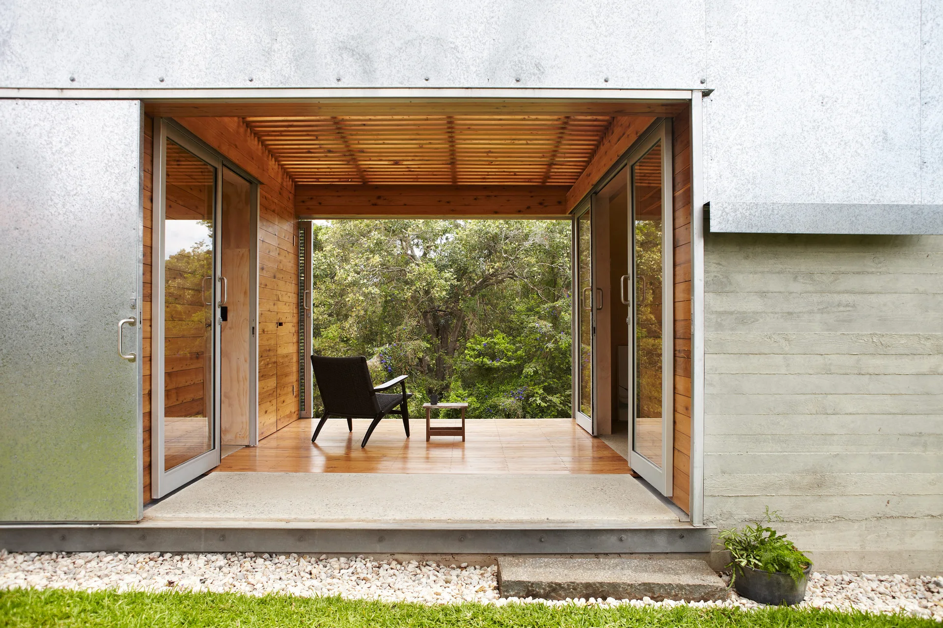 A wood-paneled covered patio features a black woven lounge chair and small side table facing a lush green forest through open sliding glass doors.