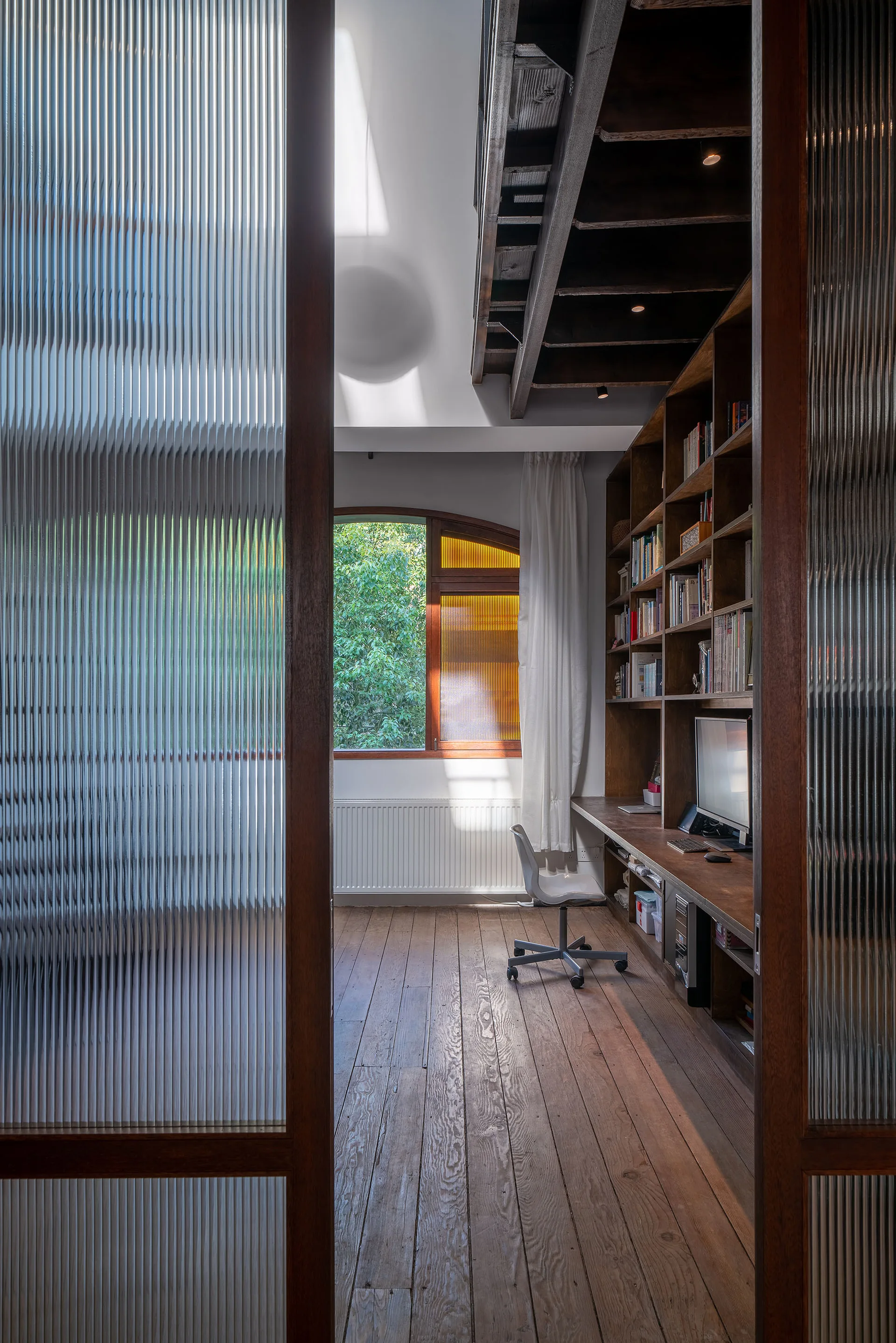 View through reeded glass sliding doors of a home office featuring built-in wooden shelving, a desk, wood floors, and an arched window.