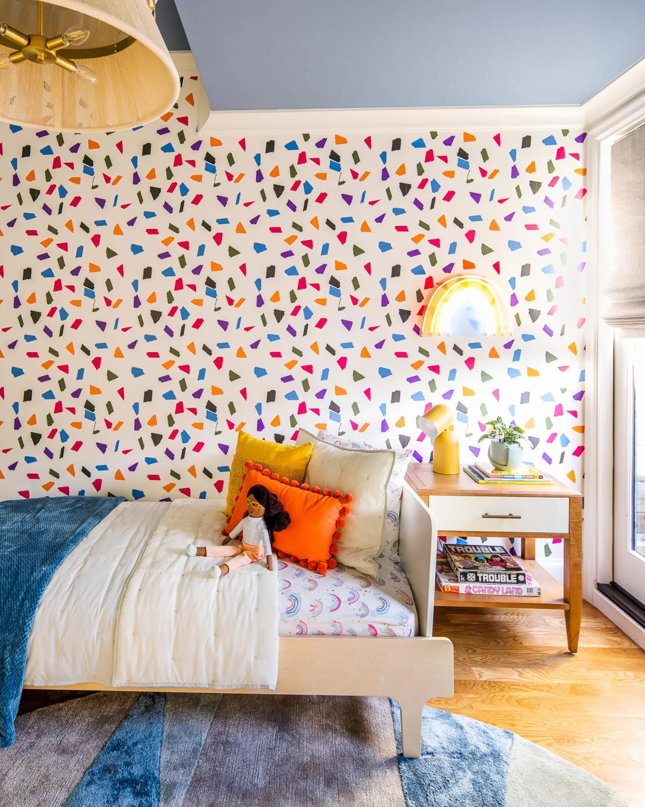 A kids bedroom features multi-colored confetti wallpaper, a light wood bed with plush pillows, a wooden nightstand, and a blue painted ceiling.