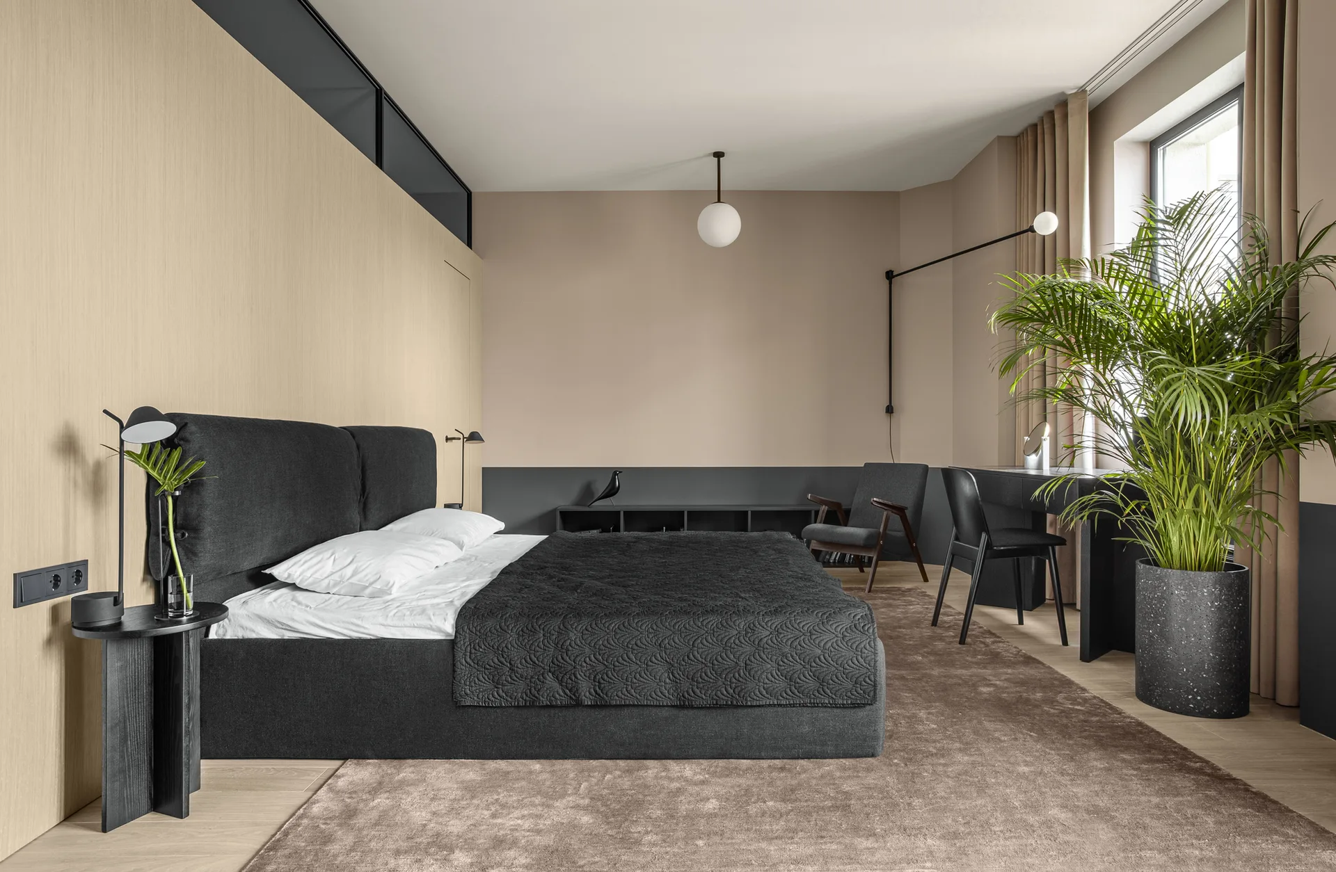 A contemporary bedroom features an upholstered charcoal bed, light wood wall paneling, a large potted palm, and a desk under a window.