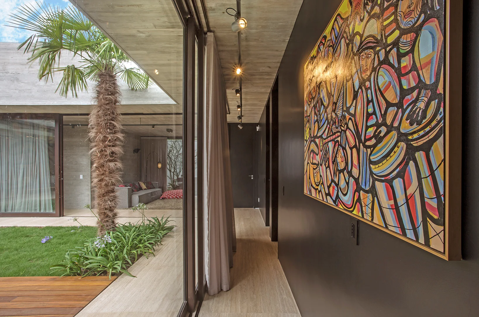 A hallway featuring a dark wall with a large colorful painting, light stone flooring, and a glass wall opening onto an outdoor courtyard with a palm tree.