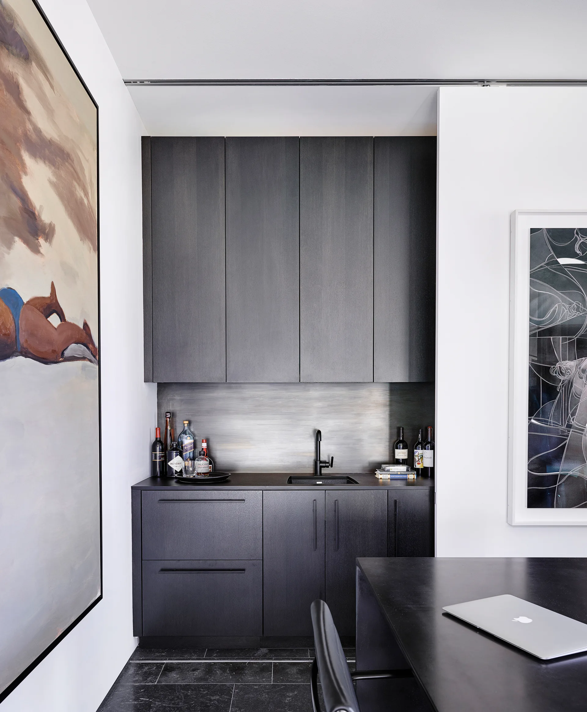A modern wet bar features matte black cabinets, an integrated sink, a metallic backsplash, and large artwork on the adjacent white walls.