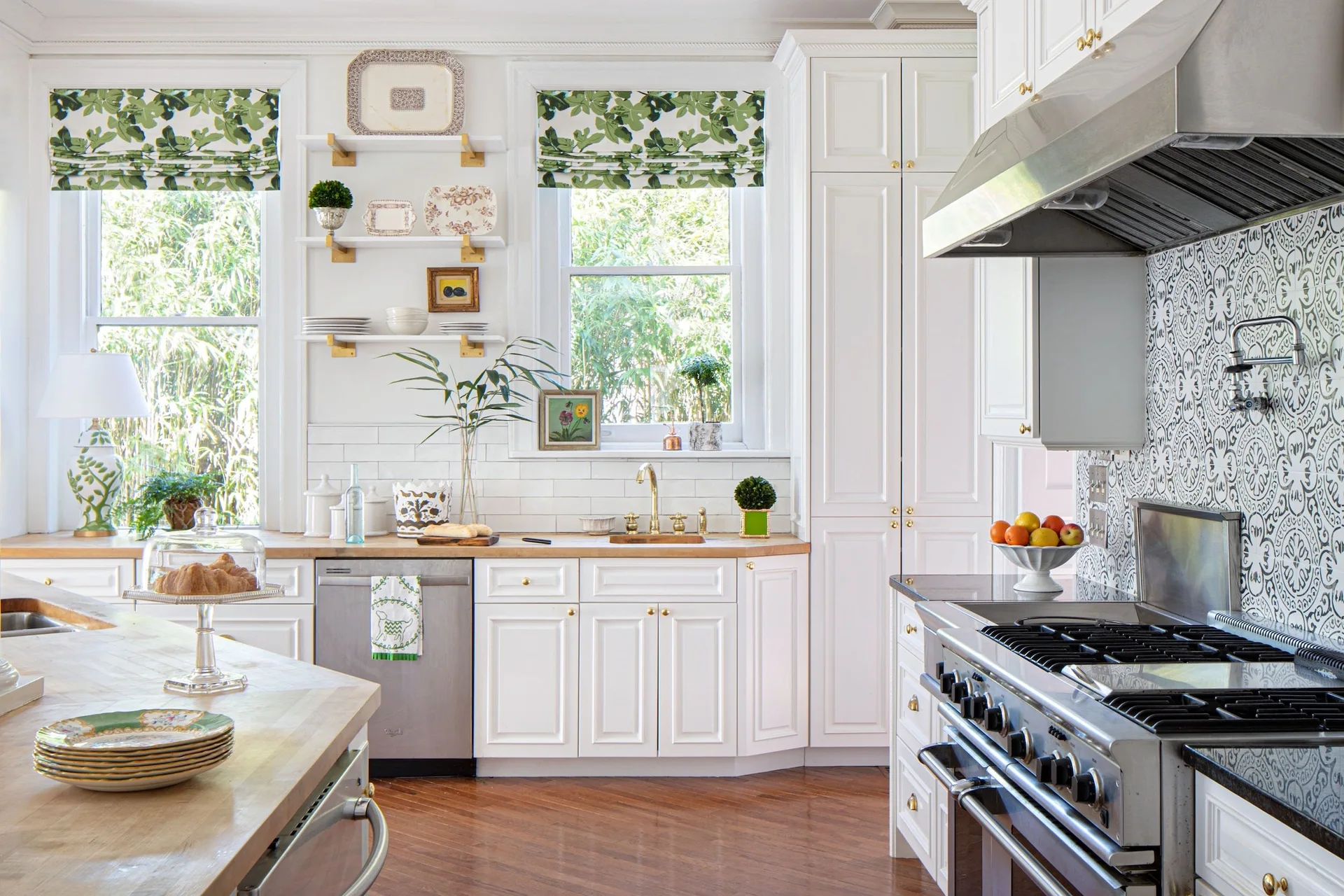 A white kitchen featuring custom wood countertops, a patterned tile backsplash, and green leafy Roman shades over the windows.