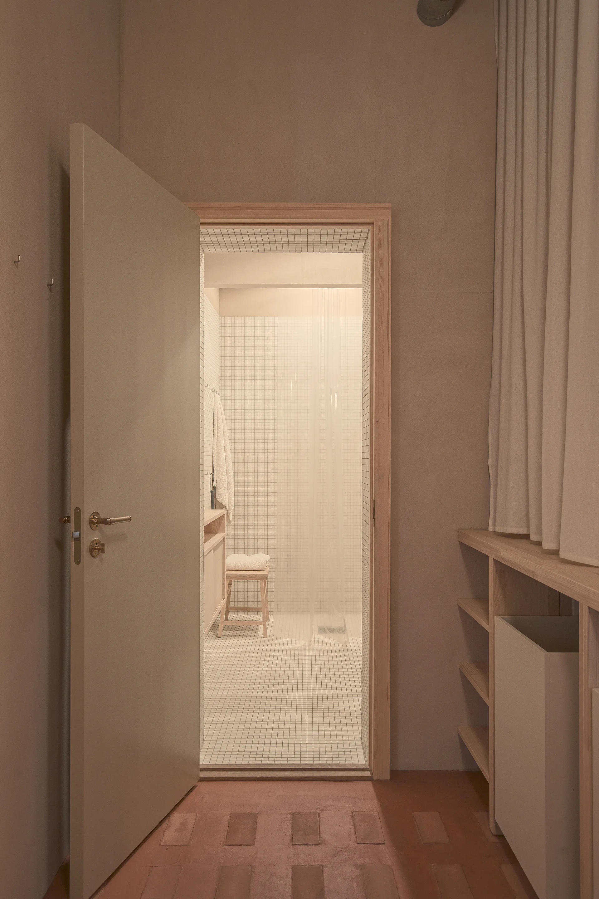An open neutral-toned door reveals a minimalist bathroom featuring small white grid mosaic tiles, a light wood stool, and warm lighting.