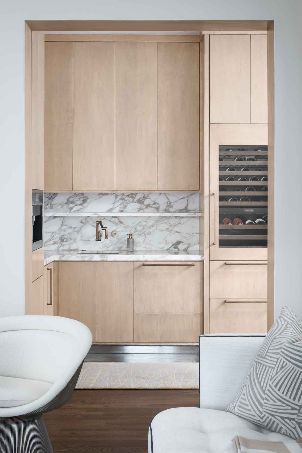 A built-in light wood home bar features a veined marble slab backsplash, an integrated wine cooler, and a small sink nestled inside a custom alcove.